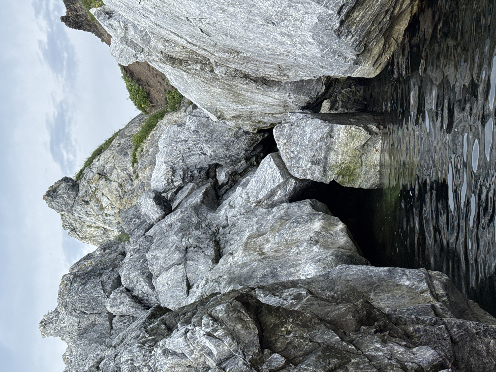 The image shows a rocky shoreline with large, grey boulders. The water is dark and appears calm with ripples on the surface. There is a dark opening between two large rocks, suggesting a cave or crevice. The sky is overcast with grey clouds. The scene is natural and rugged, with vegetation growing in some of the crevices and on the higher rocky areas. There are no people visible in the image.
