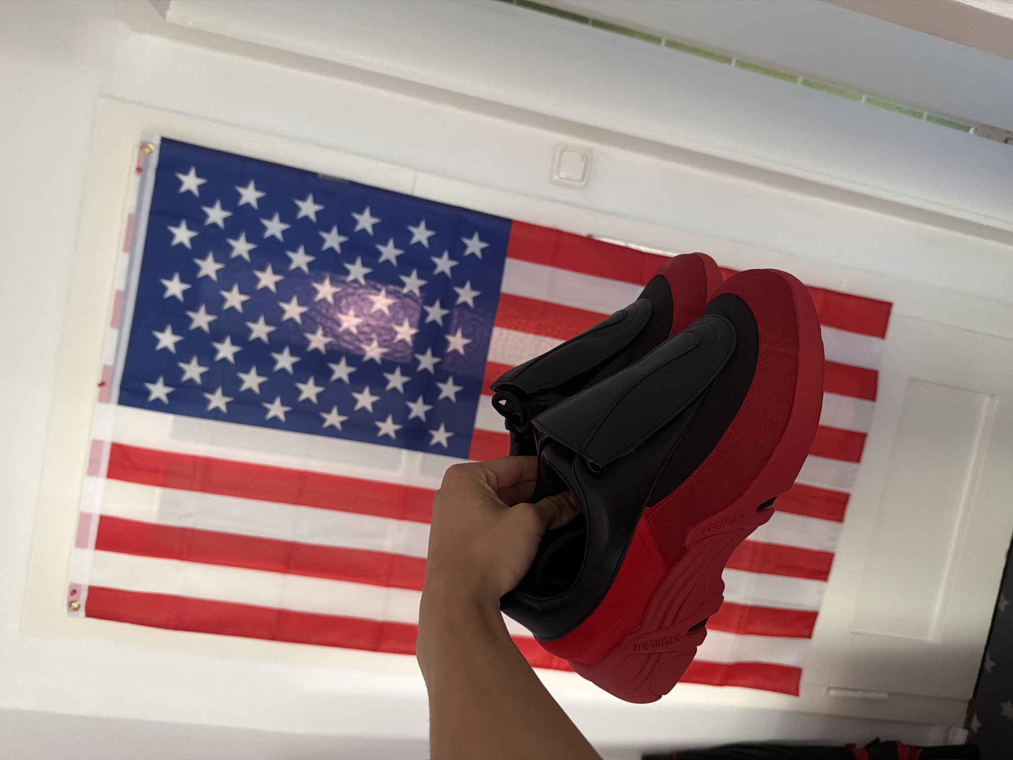 A hand holds a pair of black and red athletic shoes in front of an American flag displayed indoors. The shoes have a chunky red sole and a black upper with a black overlay. The American flag is hung vertically, with stars visible on a blue field and red and white stripes. The background appears to be a white wall with vertical blinds. The image is a medium shot, with the shoes and flag occupying the majority of the frame. The location is unknown, but it is not in Nienburg/Weser, Germany.