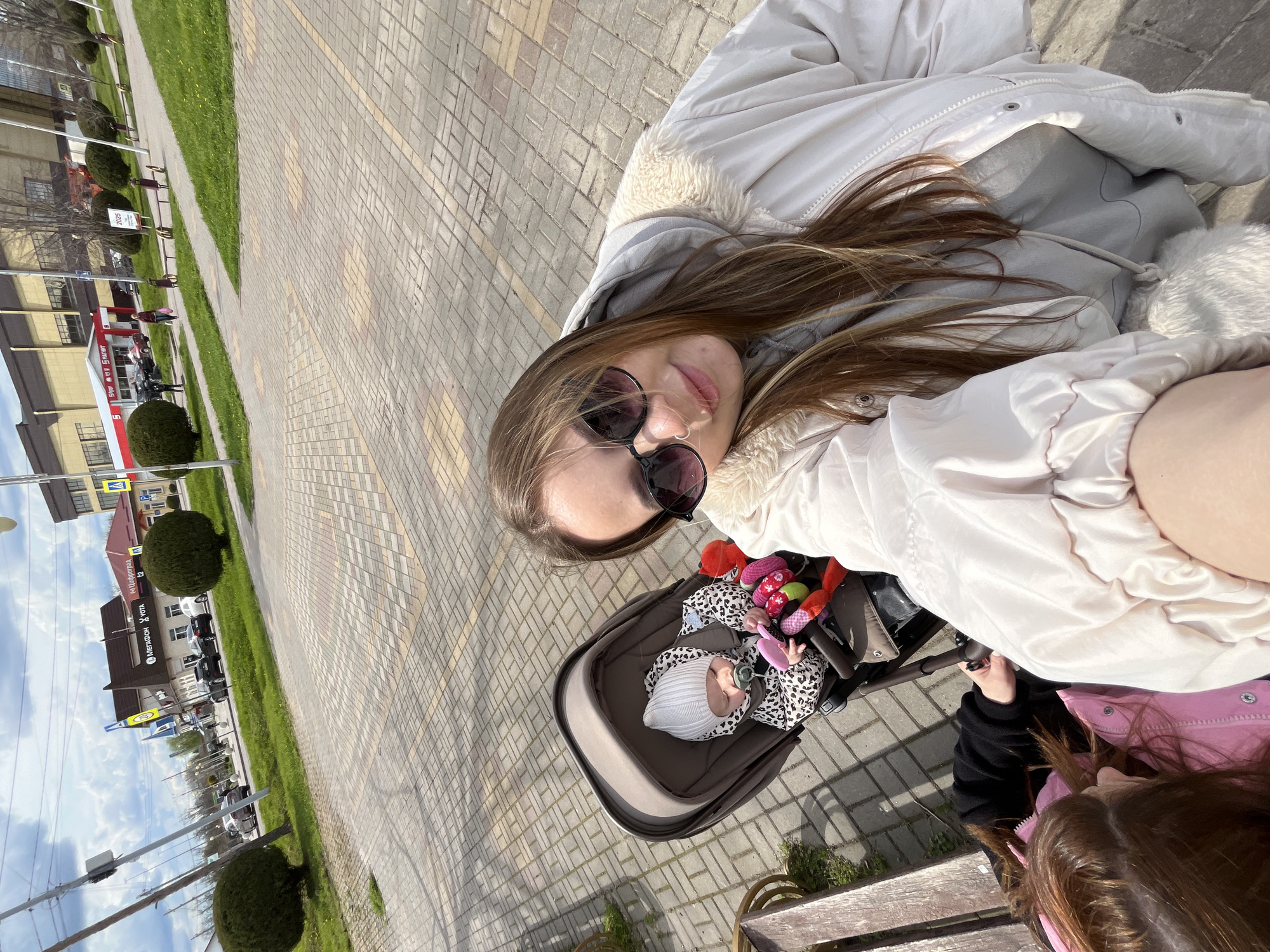 The image shows a selfie of a woman wearing sunglasses and a white jacket, presumably in the city of Novopokrovskaya, Russia, due to the presence of a store or establishment with a sign in Russian. A baby in a stroller is also visible in the frame, along with another child with brown hair wearing a pink jacket. They are on a sidewalk with brick pavement, next to a grassy area with trimmed bushes. Buildings are visible in the background.