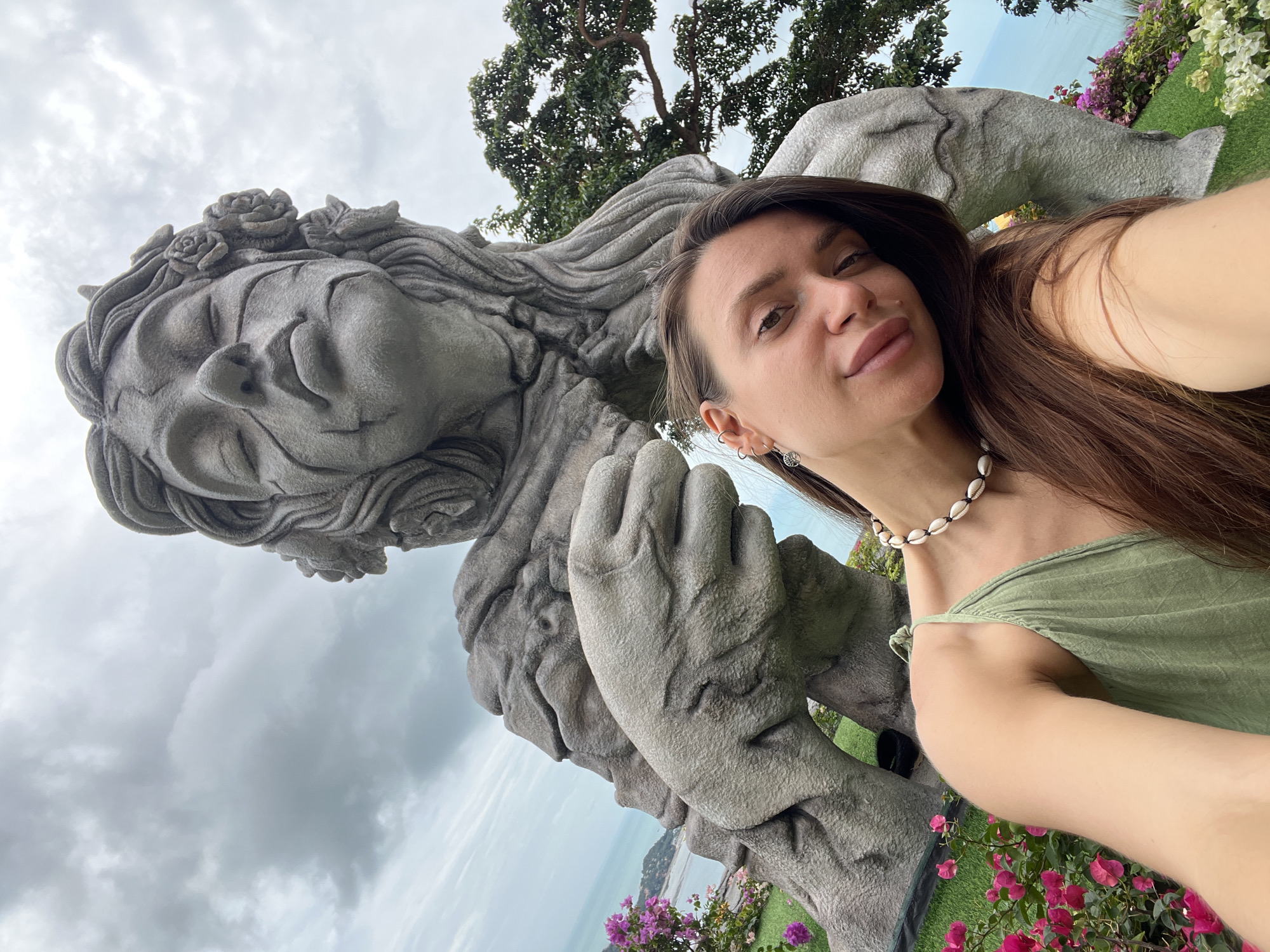 A woman is taking a selfie in front of a large, ornate stone sculpture. The woman is wearing a green top and a shell necklace. The sculpture is a reclining figure with flowers in its hair. The background includes trees, flowers, and a view of the water and coastline. The location is in the city of Unknown, Unknown.
