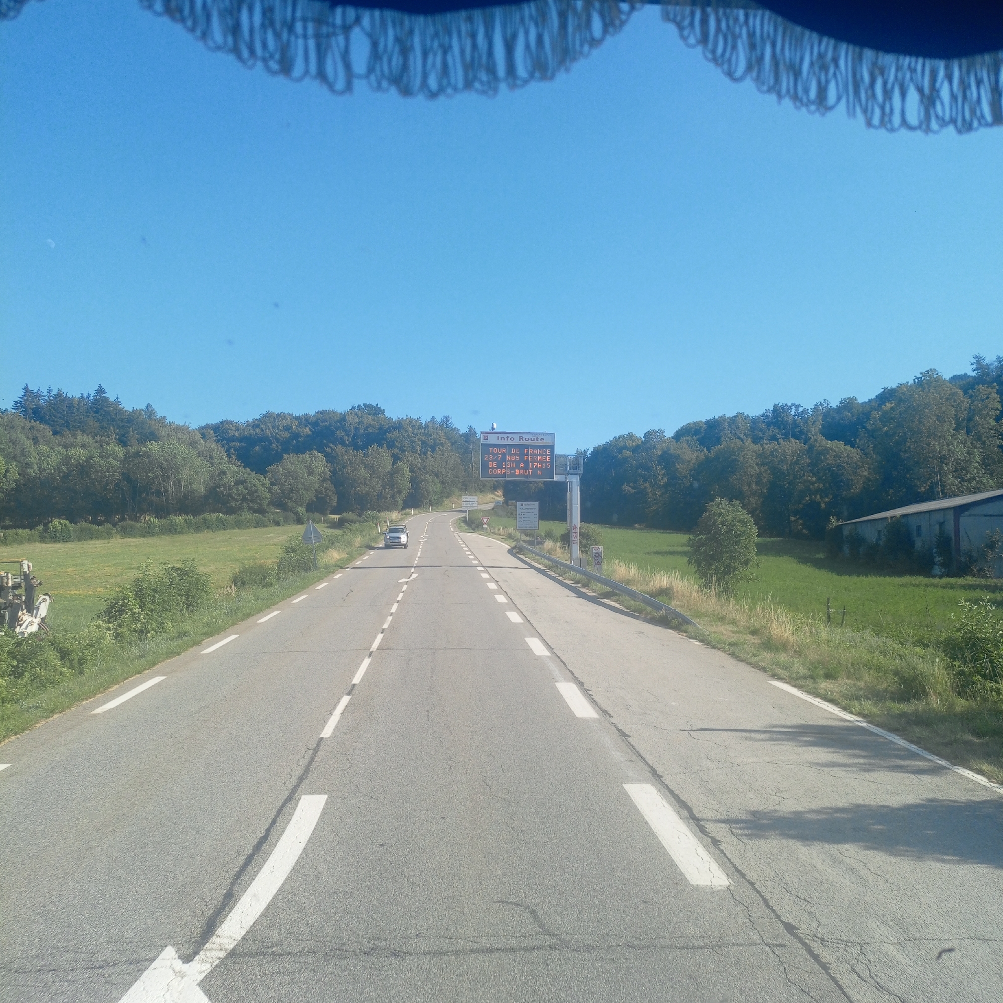 The image shows a view from a vehicle driving on a rural road in Laye, France. The road is paved with white dashed lines indicating lanes. On the right side of the road, there is a grass field and trees. Further ahead, there is a large electronic sign displaying "INFO ROUTE TOUR DE FRANCE 23/7 N85 FERMEE DE 13H A 17H CORPS-BRUT N". This indicates that Route N85 is closed due to the Tour de France on July 23rd from 1 PM to 5 PM. A white car is visible driving in the opposite direction on the road. The sky is clear and blue. There is a structure with a roof and fringe visible at the top of the image, suggesting it's the interior of a vehicle or a canopy.