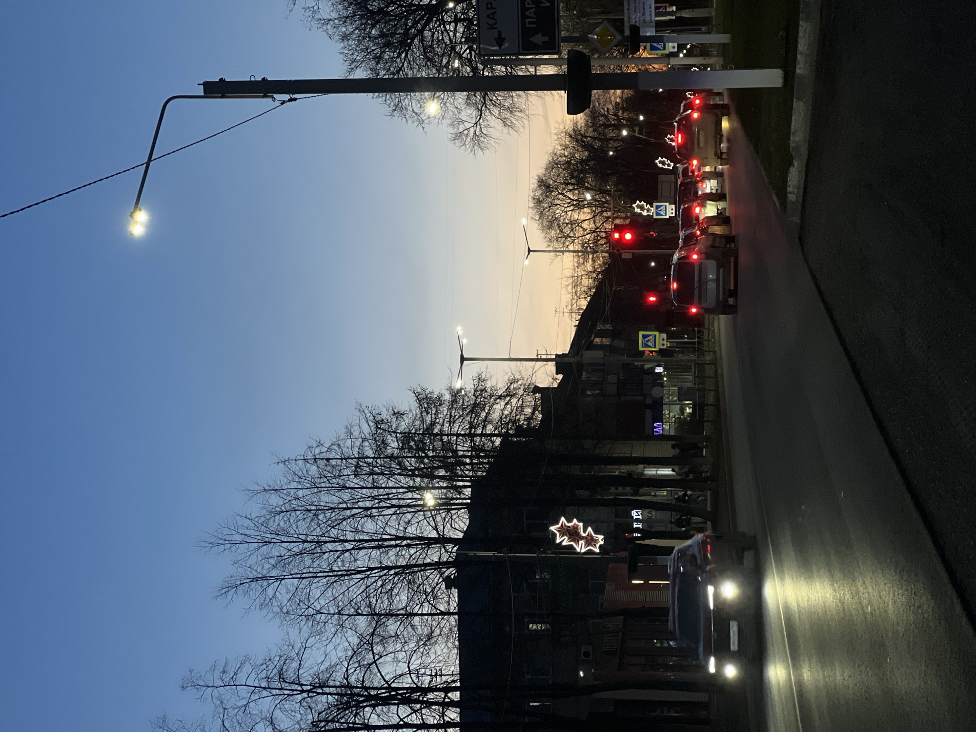 The image depicts a street scene in Chaikovsky, Russia, at twilight. The sky transitions from blue to orange. Traffic is stopped at a red light, with several cars visible, including a car with illuminated headlights facing the camera. A street lamp illuminates the left side of the image. Bare trees line the street on both sides, and buildings are visible in the background. Christmas star decorations are hanging. Street signs are also visible.