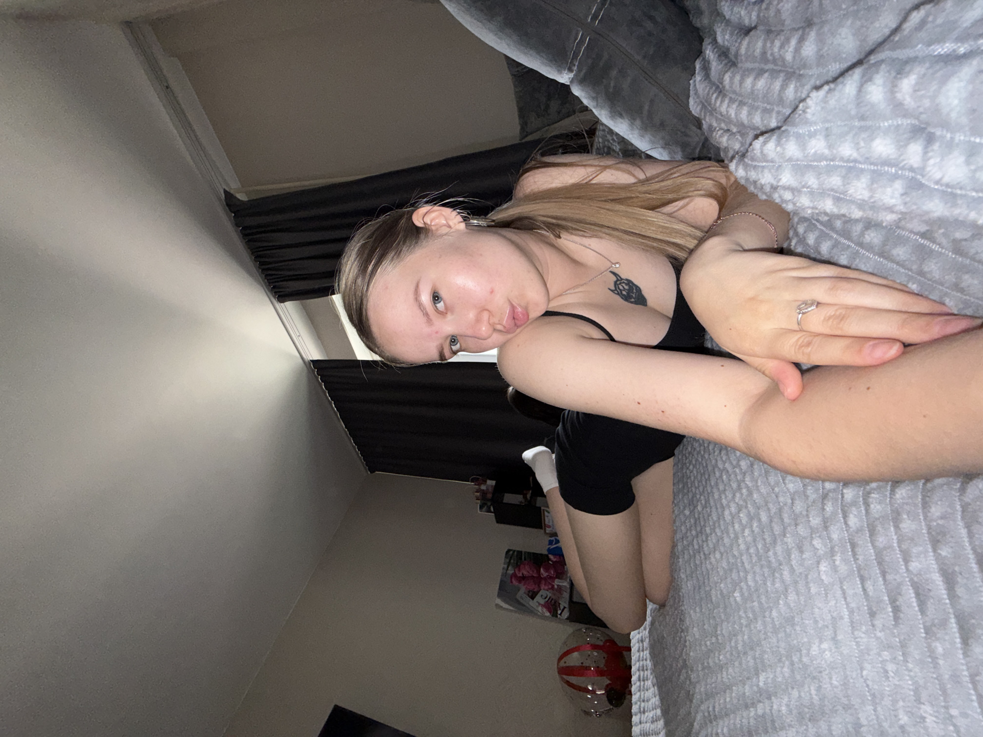 A young woman is lying on a grey couch or bed in what appears to be a bedroom. She is fair-skinned, has blonde hair pulled back in a ponytail, and is wearing a black top. She has a tattoo on her chest and a ring on her finger. The background is dimly lit, showing a dark curtain and some items on the floor, including a balloon and magazines. The location is unknown, but it is not possible to determine if it is in Chaikovsky, Russia.