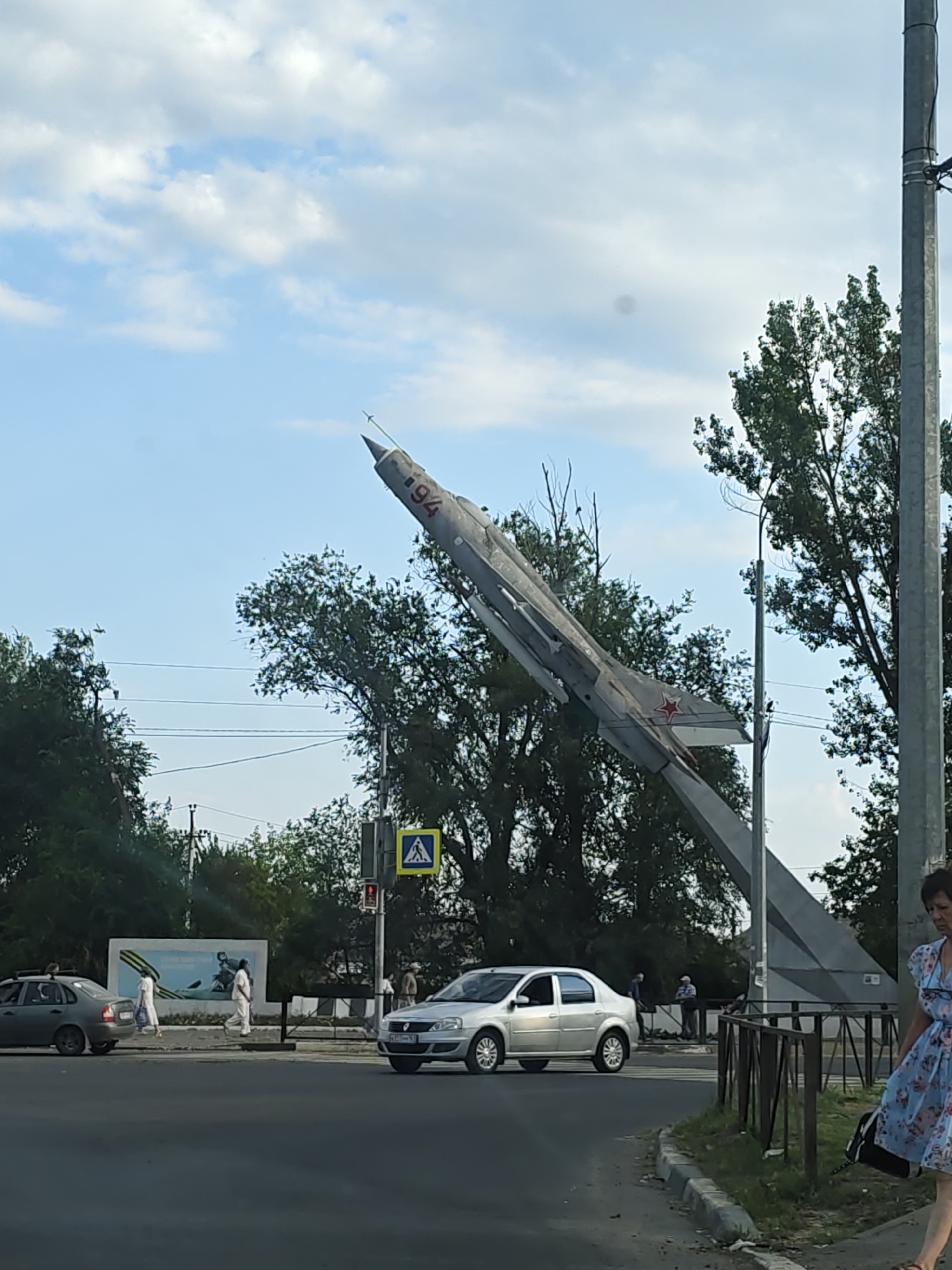 The image shows a monument of a MiG-21 fighter jet in Novocherkassk, Russia. The jet is mounted on a concrete structure and points upwards. In the background, there are trees and a cloudy sky. A street runs in the foreground with several cars and pedestrians. A woman in a floral dress is walking on the right side of the image. A billboard with an image of a war scene is visible behind the street. The scene depicts a monument dedicated to aviation, likely commemorating military aviation.