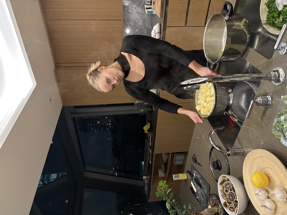 A woman wearing a black top is in a kitchen. She is pouring potatoes from a pot into a colander in the sink. Steam is rising from the potatoes. A large pot sits next to the colander. A cutting board, a bowl with dried lemons, a lemon, and garlic bulbs are on the countertop. A window with a city view is visible in the background.