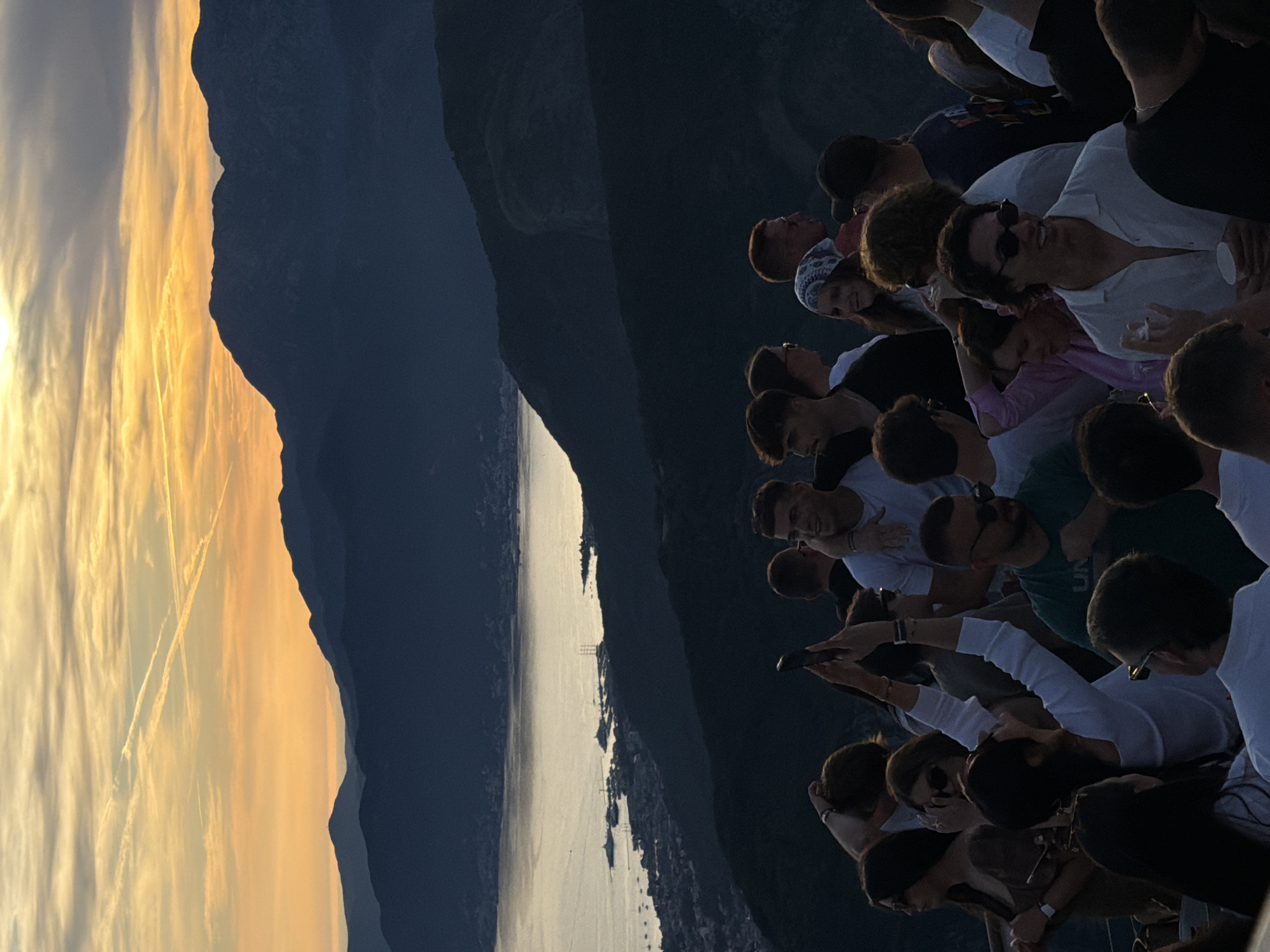 A group of people stand on a mountaintop overlooking a bay at sunset. The sky is filled with orange and yellow hues from the setting sun, casting a warm glow over the scene. Silhouetted mountains rise in the background, with a town and boats visible on the coast below. The people in the foreground are casually dressed, some holding phones and drinks. The overall atmosphere appears relaxed and social, with a sense of camaraderie amongst the group. The image captures a beautiful moment of natural beauty and human connection.