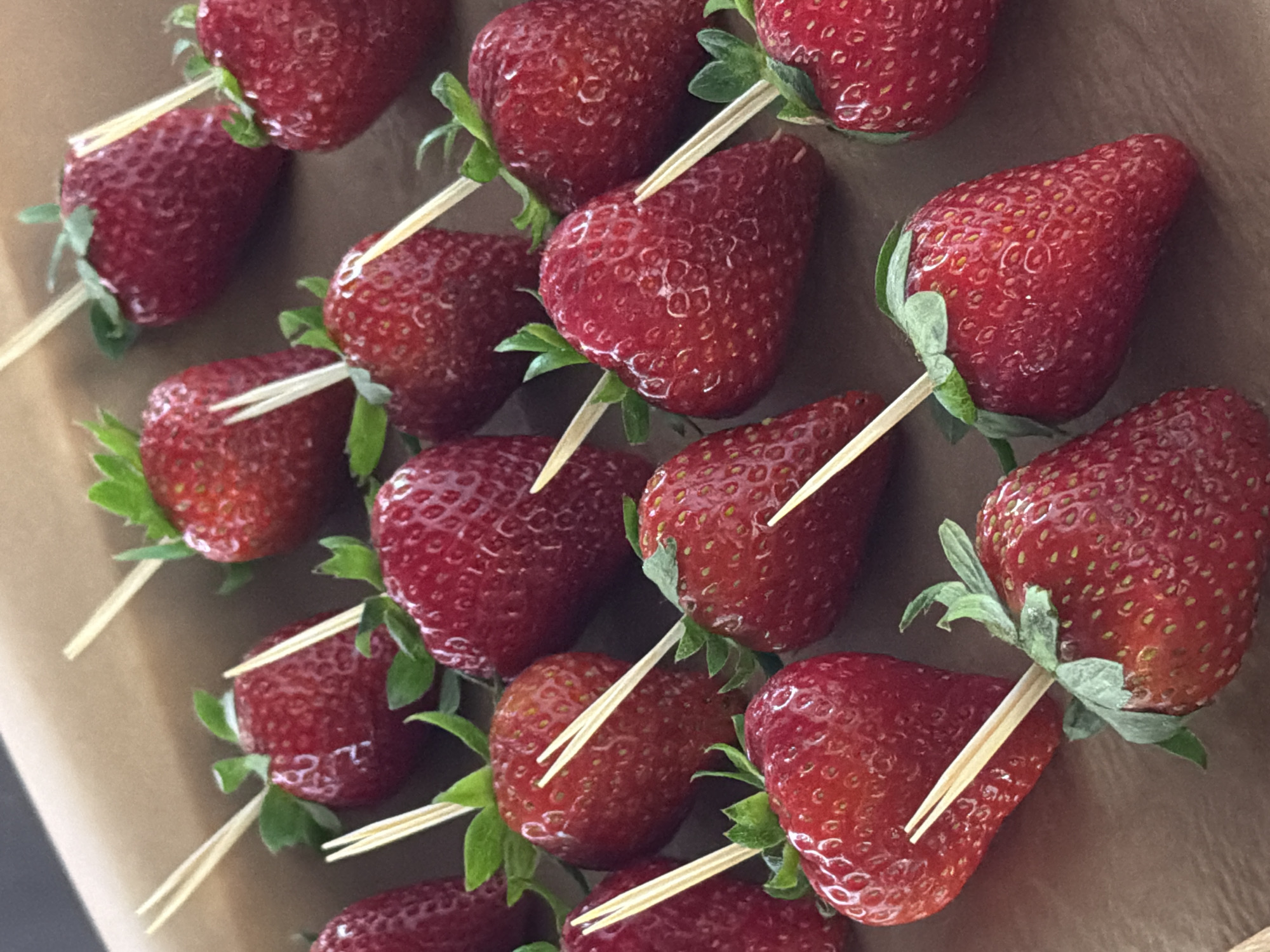 This media file shows a close-up, overhead view of a collection of strawberries skewered on toothpicks, arranged on a brown surface, likely a table or tray. The strawberries are a vibrant red, with visible seeds and green leafy tops. Some of the strawberries are partially obscured by others, indicating they are densely packed. The toothpicks are light-colored and pointed, inserted into the fruit to hold them in place. The background is a plain, matte brown color, suggesting an indoor setting. There are no people visible, and the focus is entirely on the fruit. The lighting appears to be even, with no harsh shadows, hinting at a well-lit indoor environment. The overall impression is of a prepared snack or appetizer, possibly for a party or event. There is no visible text in the image. The context provided, "Saint Petersburg, Russia," offers a geographical location but does not directly inform the visual content beyond the general possibility of strawberries being available there.