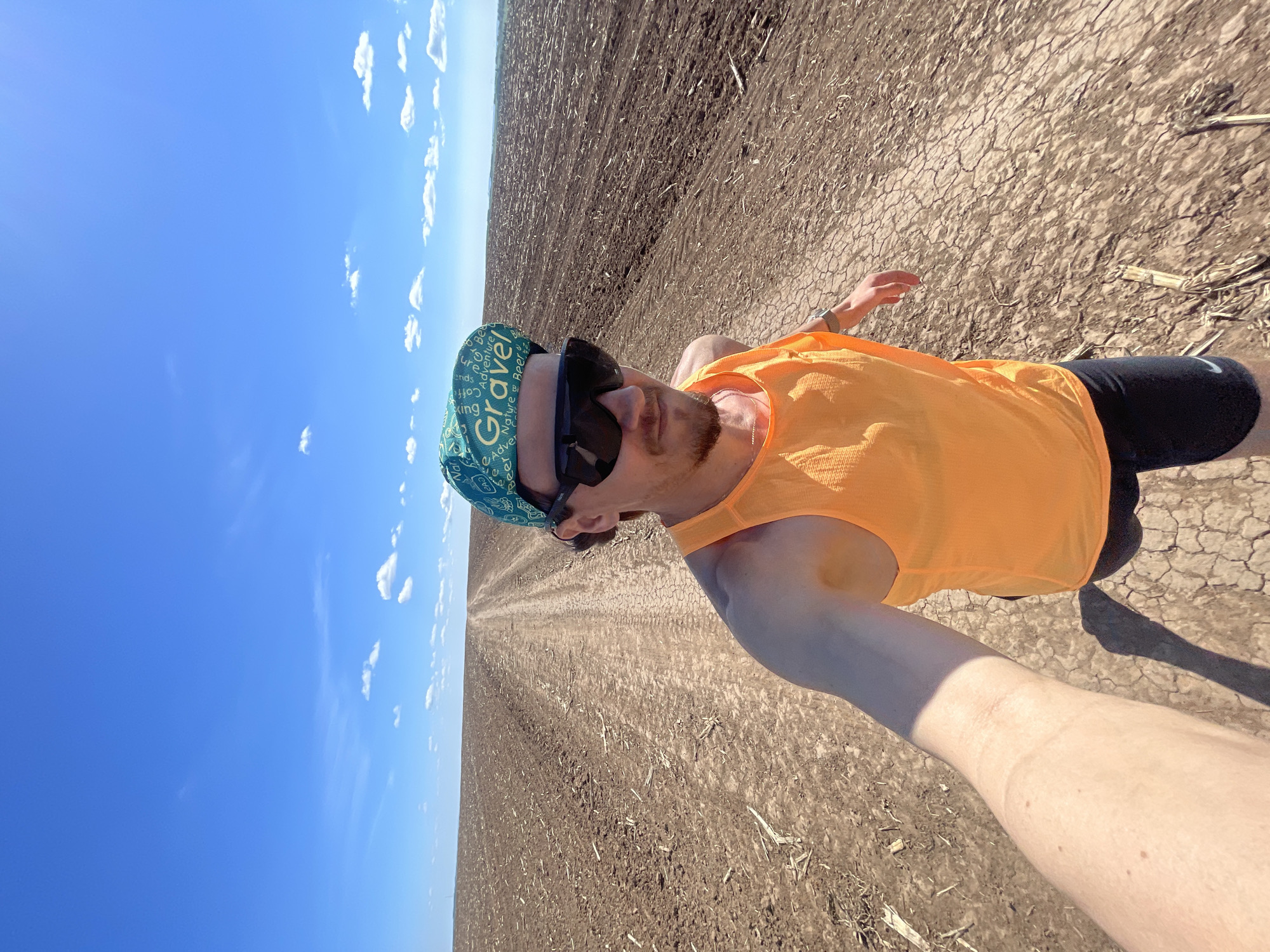 A man takes a selfie outdoors in a rural area. He is wearing a blue and green cycling cap, sunglasses, an orange tank top, and black shorts. He is standing on a dry, cracked dirt surface, possibly a field or a dirt road. In the background, there is a vast plowed field stretching to the horizon, with a clear blue sky dotted with a few clouds above. The location may be near the city of Perevoznoe, Russia.