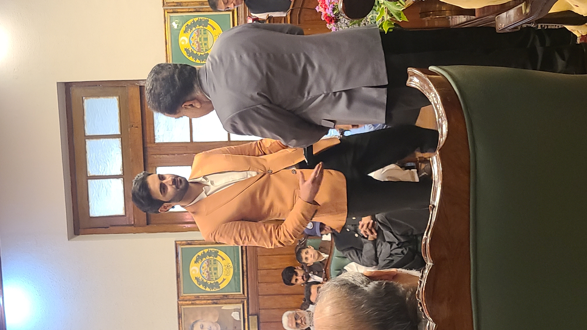 A medium indoor shot taken in Quetta City Tehsil, Pakistan, depicts a man in a tan blazer gesturing with his left hand while facing another man in a gray blazer. A window with panes is behind the man in the tan blazer, and framed emblems adorn the wall. A crowd of people, some seated, are in the foreground. A green upholstered chair frame occupies the right of the image.