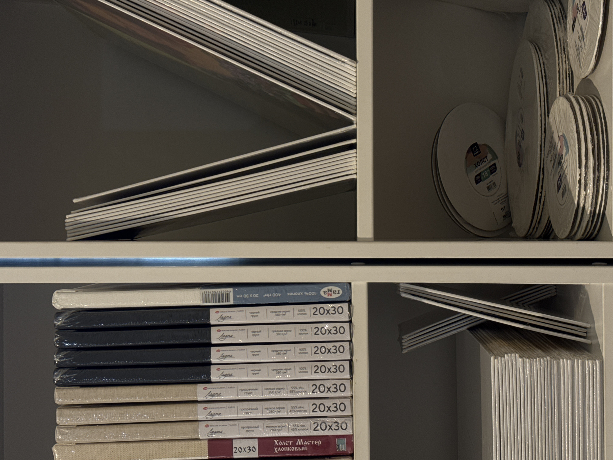 The image shows a close-up, eye-level, vertical shot of shelves stocked with art supplies in what appears to be a retail environment. The shelves are white and are divided into two sections by a vertical support. The left section of the shelves contains several canvases stacked vertically, each with a label indicating dimensions such as "20x30" and some text in Russian including "Холст Мастер Хлопковый" and "про грунтованный грунт". The right section of the shelves displays several large, thin art boards or canvases stacked in a fanned arrangement, leaning against each other. Below these, the left side of the bottom shelf shows a stack of square or rectangular art boards, also wrapped in plastic. The right side of the bottom shelf displays several round canvases, stacked in varying heights, with labels visible. The overall lighting is dim, creating shadows and highlighting the textures of the materials. There are no people visible in the image. The context of Moscow, Russia is not discernible from the visual information provided in the image itself.