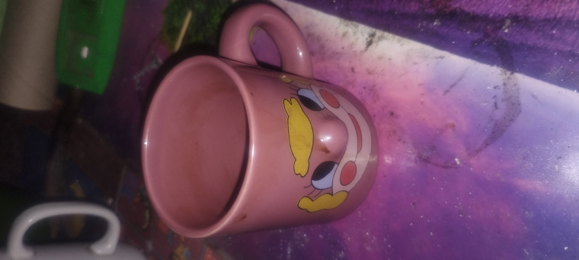 The image shows a pink ceramic mug with a cartoon face design, including blonde hair, big black eyes, red cheeks, and a smiling mouth. The mug has a handle on the right side. It is placed on a surface that appears to be a table or desk with a purple and pink speckled pattern. In the background, a white mug handle is partially visible on the left, and a green object is visible on the right. The scene is indoors, and there is no specific indication of its location within the city of Zaporizhzhia, Ukraine.