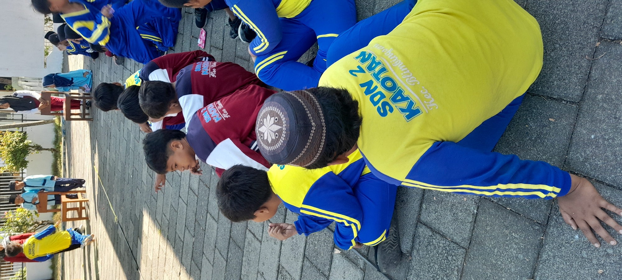 A group of young Indonesian children, likely students, are sitting on a paved area. The children are wearing various school uniforms, some in blue and yellow, others in blue and red. One child in the foreground is wearing a yellow long-sleeved shirt with "SDN 2 KAOTAN" and "Kec. Blimbingan" printed on the back. Another child in the foreground is wearing a dark patterned skullcap. In the background, there are several more children and adults. Some children are standing near chairs and a small wooden structure, suggesting an outdoor school activity or event. The setting appears to be a schoolyard or public space in Rogojampi, Indonesia, with a tiled ground and some trees visible. The lighting suggests it is daytime, possibly late morning or early afternoon.