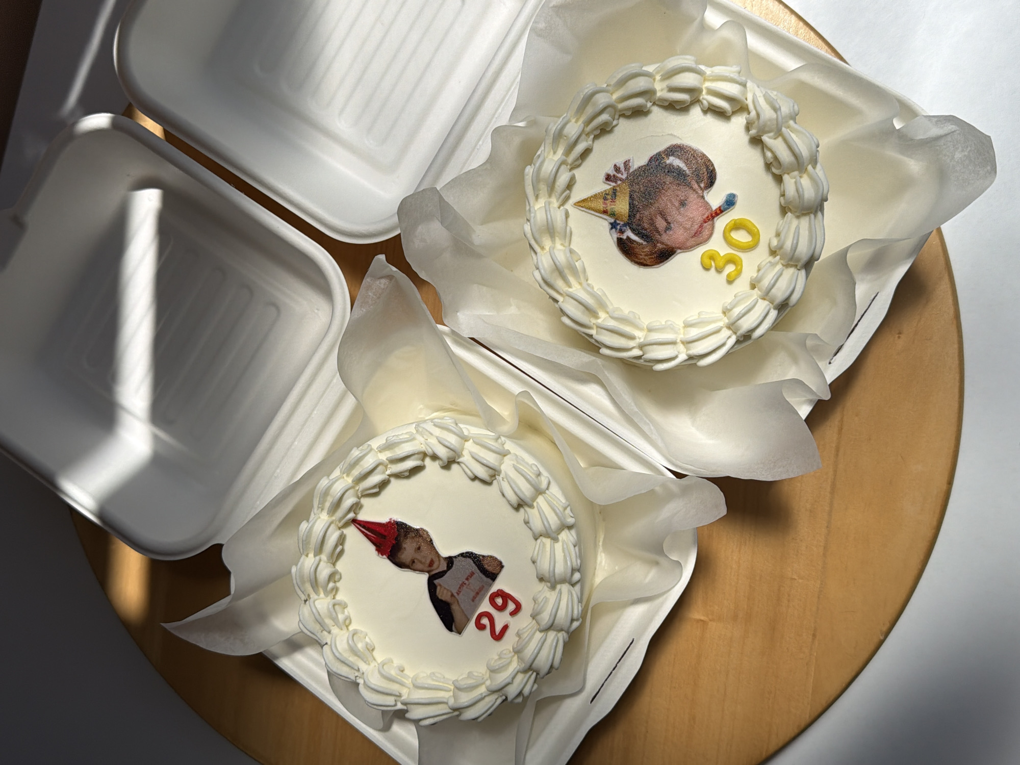 Two white cakes are visible, each decorated with a photo of a person and a number representing an age. The cake on the bottom left features a person wearing a red party hat and the number "29" in red. The cake on the top right features a different person wearing a gold party hat with a blue and red decoration, and the number "30" in yellow. Each cake has white frosting around the edges and is set on white tissue paper inside a white clamshell container. An empty clamshell container is visible on the left. The cakes are placed on a light brown, possibly wooden, surface. There is a harsh light from the left, possibly sunlight, casting shadows on the objects. This image does not give any clues about location in the city of Tel-Aviv, Israel.