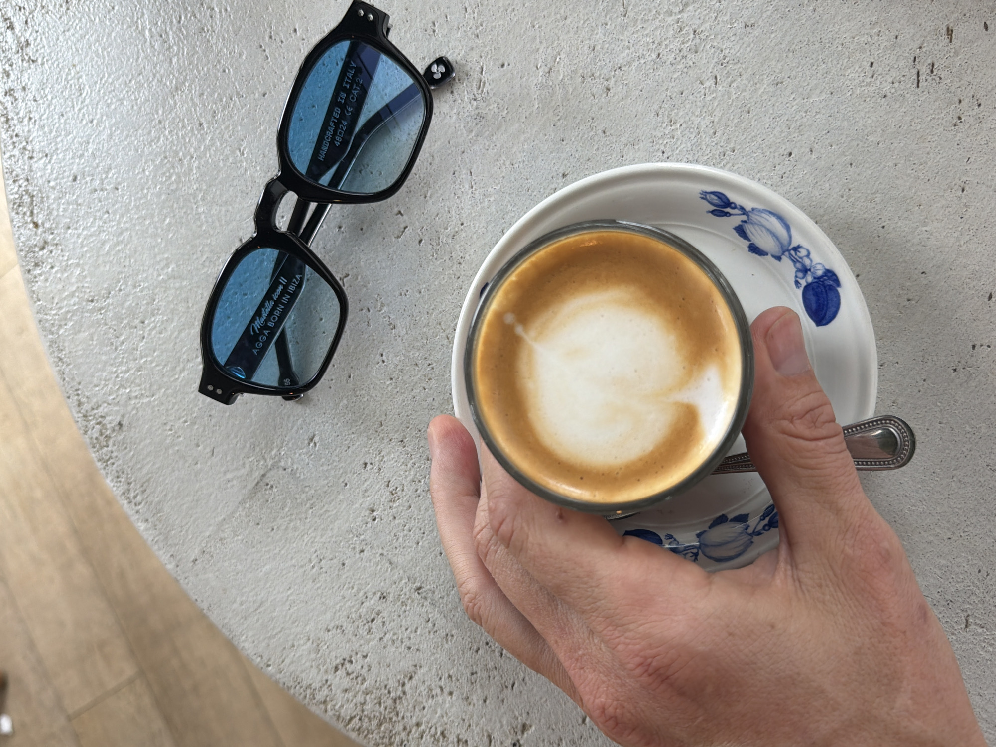An overhead shot shows a hand holding a cup of coffee with latte art on a saucer with blue floral accents. The cup and saucer sit on a textured, light-colored surface next to a pair of black-framed sunglasses with blue lenses. The sunglasses are placed on the surface with the lenses facing upwards. Text on the sunglasses reads: "Mustella icon II AGGA BORN IN IBIZA" and "HANDCRAFTED IN ITALY 48024 CE CAT.2". The background shows a light-colored tiled floor. The image appears to be taken in Miami Beach, Florida, United States of America.