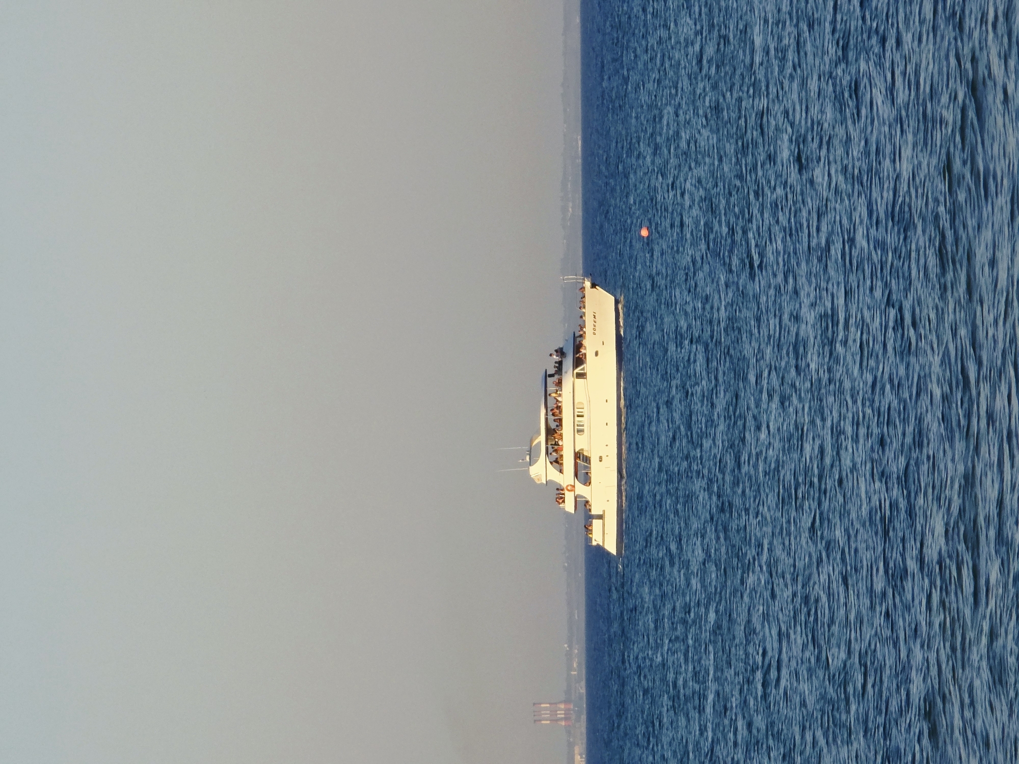 A white multi-deck passenger boat, labeled "DOREMI," is cruising on the blue sea under a clear, hazy sky. The boat is populated with numerous passengers standing on its decks, some appearing to be seated. In the distance, the Larnaca coastline is visible, featuring industrial smokestacks and low-lying structures. A small orange buoy floats on the water's surface to the right of the boat. The sea exhibits moderate ripples.