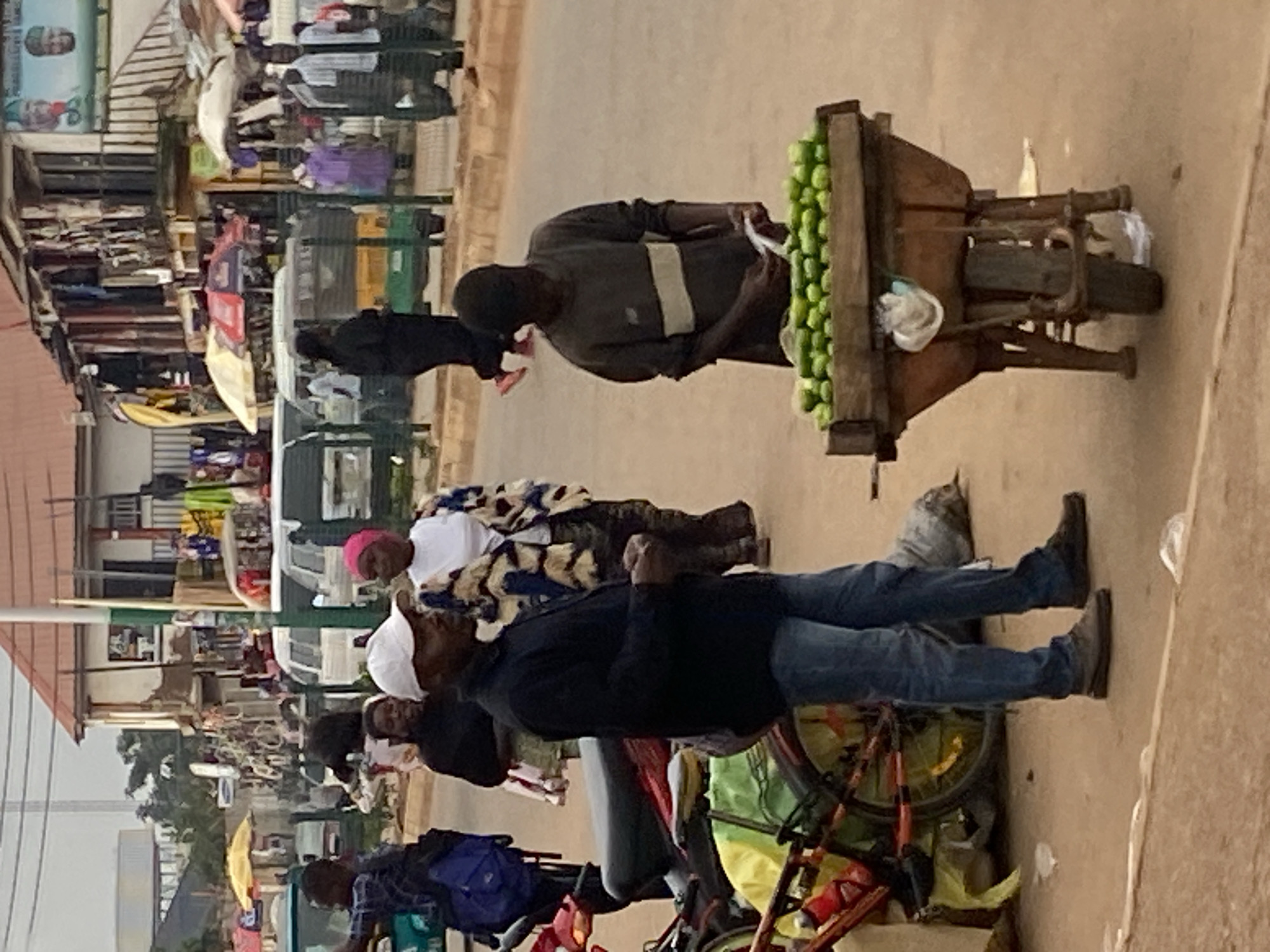 The image captures a street scene in Bukuru, Nigeria, featuring a bustling market environment. In the foreground, a man in a dark jacket and blue jeans stands to the left, facing right, his hands clasped. To his right, another individual, possibly a young man, is tending to a wooden wheelbarrow filled with green produce, appearing to be limes or cucumbers. He is dressed in a grey long-sleeved shirt and is looking down at the produce. Behind them, a variety of market stalls with awnings and goods are visible, along with several people walking and interacting. Vehicles, including what appears to be a tuk-tuk and other vans, are present in the background, suggesting active commerce and transportation. The overall atmosphere is that of a busy, outdoor marketplace in an urban setting.