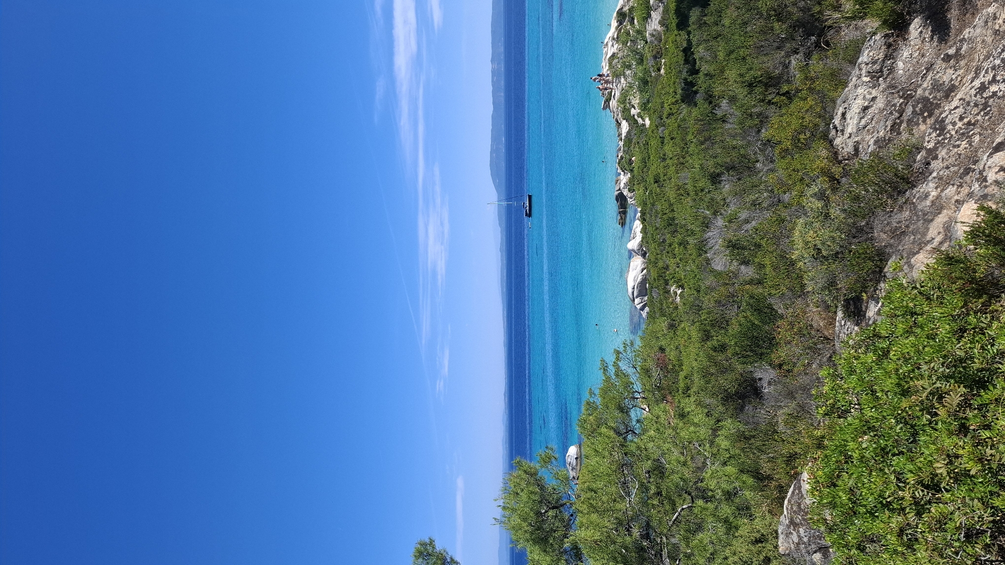 This image depicts a vibrant Mediterranean seascape on a clear, sunny day. The scene is dominated by the brilliant turquoise waters of the sea, contrasting with a deep blue sky filled with wispy white clouds. The water is so clear that the seabed and submerged rocks are visible in some areas.
Along the right side of the image, a rocky coastline is covered with lush green vegetation, including trees and shrubs. Large, white boulders are scattered among the rocks and along the water's edge. In the distance, a sailboat with a single sail is anchored in the calm water.
There are several people visible along the rocky shoreline, some appearing to be relaxing or sunbathing. A few other individuals are in the water, suggesting a leisurely day at the beach. The overall mood is one of tranquility and natural beauty, characteristic of a summer vacation spot in Greece, specifically Toroni. The lighting suggests it is daytime, likely late morning or early afternoon, given the bright sunlight and clear conditions. There is no visible text in the image.