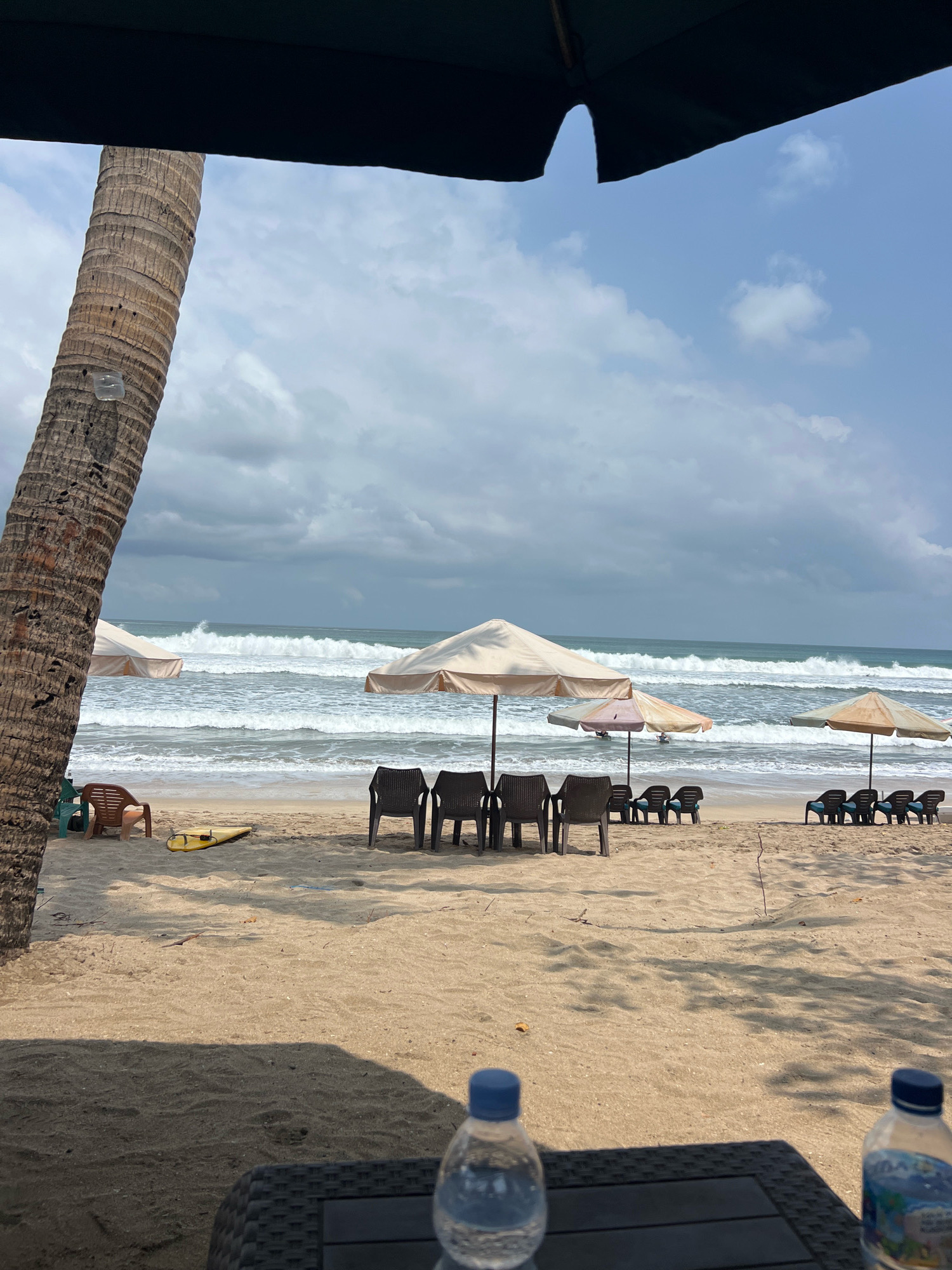 The image captures a view of Kuta Beach in Bali, Indonesia, on a cloudy day. In the foreground, a dark umbrella casts a shadow over a portion of the sandy beach. Two plastic water bottles sit on a dark woven table. A large palm tree trunk stands prominently on the left side of the frame.
Beyond the foreground, the beach is dotted with empty lounge chairs and umbrellas. A yellow surfboard rests on the sand near some chairs. The ocean is active with moderate waves crashing onto the shore, creating a dynamic seascape under a partly cloudy sky. In the distance, a few people can be seen in the water, enjoying the surf. The overall scene depicts a typical beach setting with amenities for visitors, despite the overcast weather.