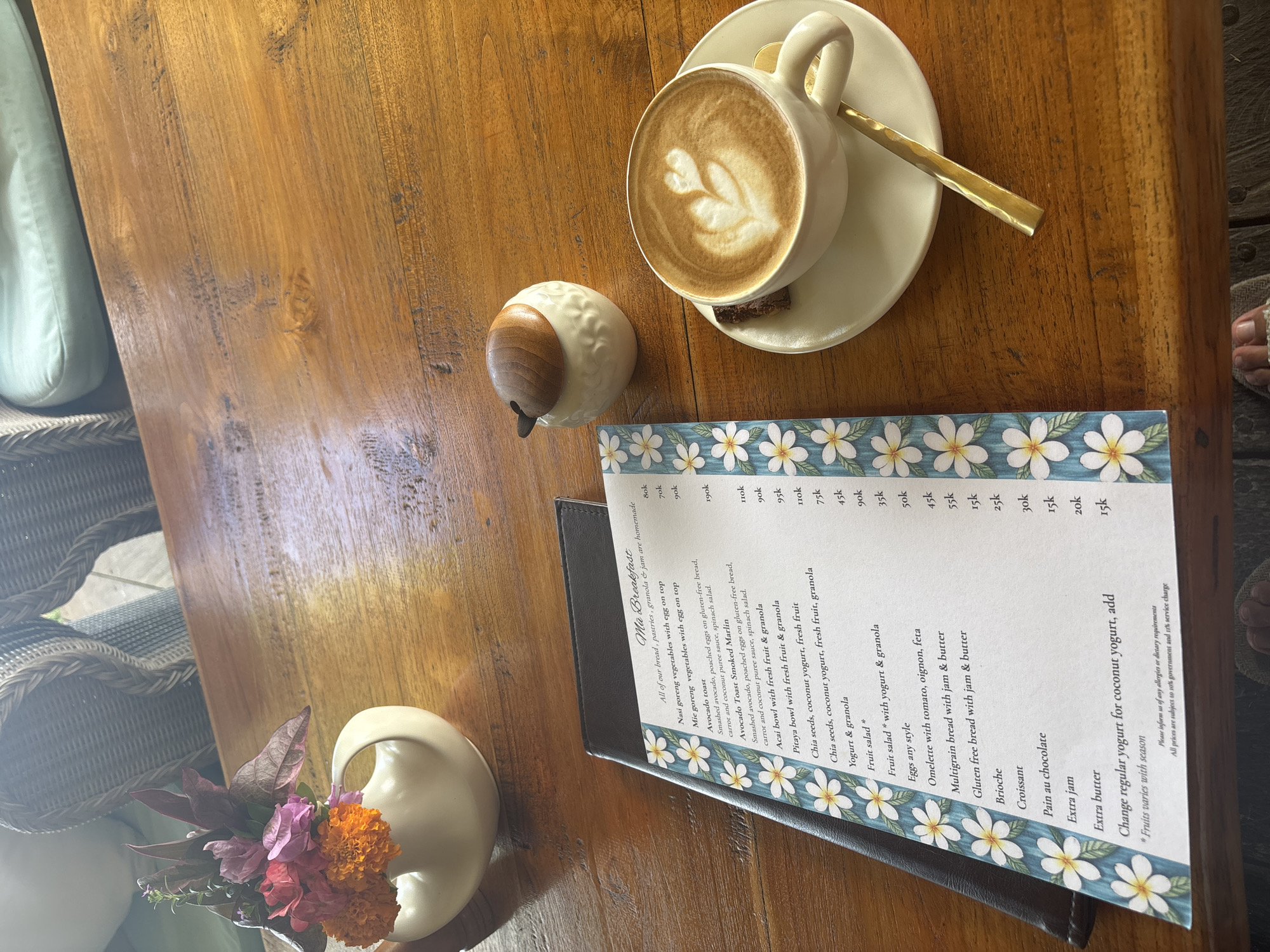 Here is a concise description of the scene:
The image shows a wooden table setting, likely in a cafe in Kuta Selatan, Indonesia. Visible on the table are: a cup of coffee with latte art, a small ceramic container (possibly for milk or sugar), a menu titled "Mia Breakfast" advertising breakfast items such as Nasi goreng, Avocado toast, Açai bowls, yogurt, omelettes, breads and jams, and flowers in a white vase. The table is partially covered by a woven chair. A foot is visible in the top right corner.