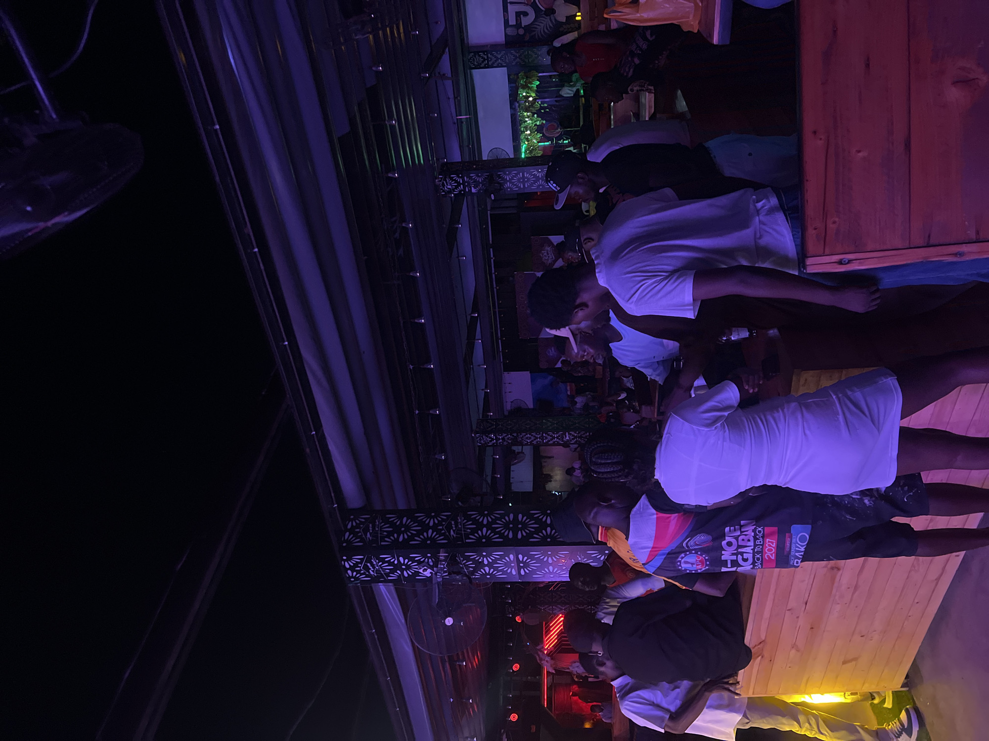 The image shows a nighttime scene in Howayi, Nigeria, likely at an outdoor bar or club. Several people are present, including some wearing light-colored clothing. Some people appear to be standing near a wooden structure, possibly a bar or seating area. Overhead, there's a metal structure with decorative elements and exposed ductwork. Lighting in the area appears to be a mix of artificial sources, including red, green, and purple lights. There is a fan hanging from the metal structure.