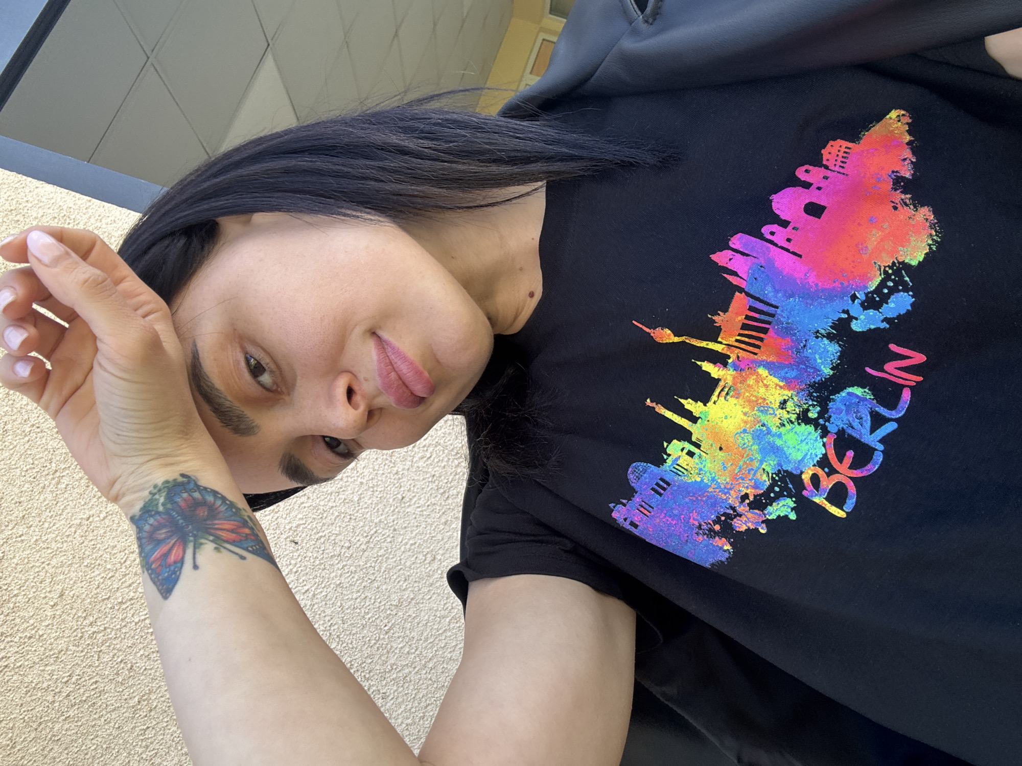 A medium shot depicts a woman with dark hair wearing a black t-shirt with a multicolored depiction of the Berlin skyline and the word "Berlin" on it. She has a butterfly tattoo on her left wrist. The background appears to be a textured wall, possibly outdoors. The location is unknown, and there is no indication of Tbilisi, Georgia.