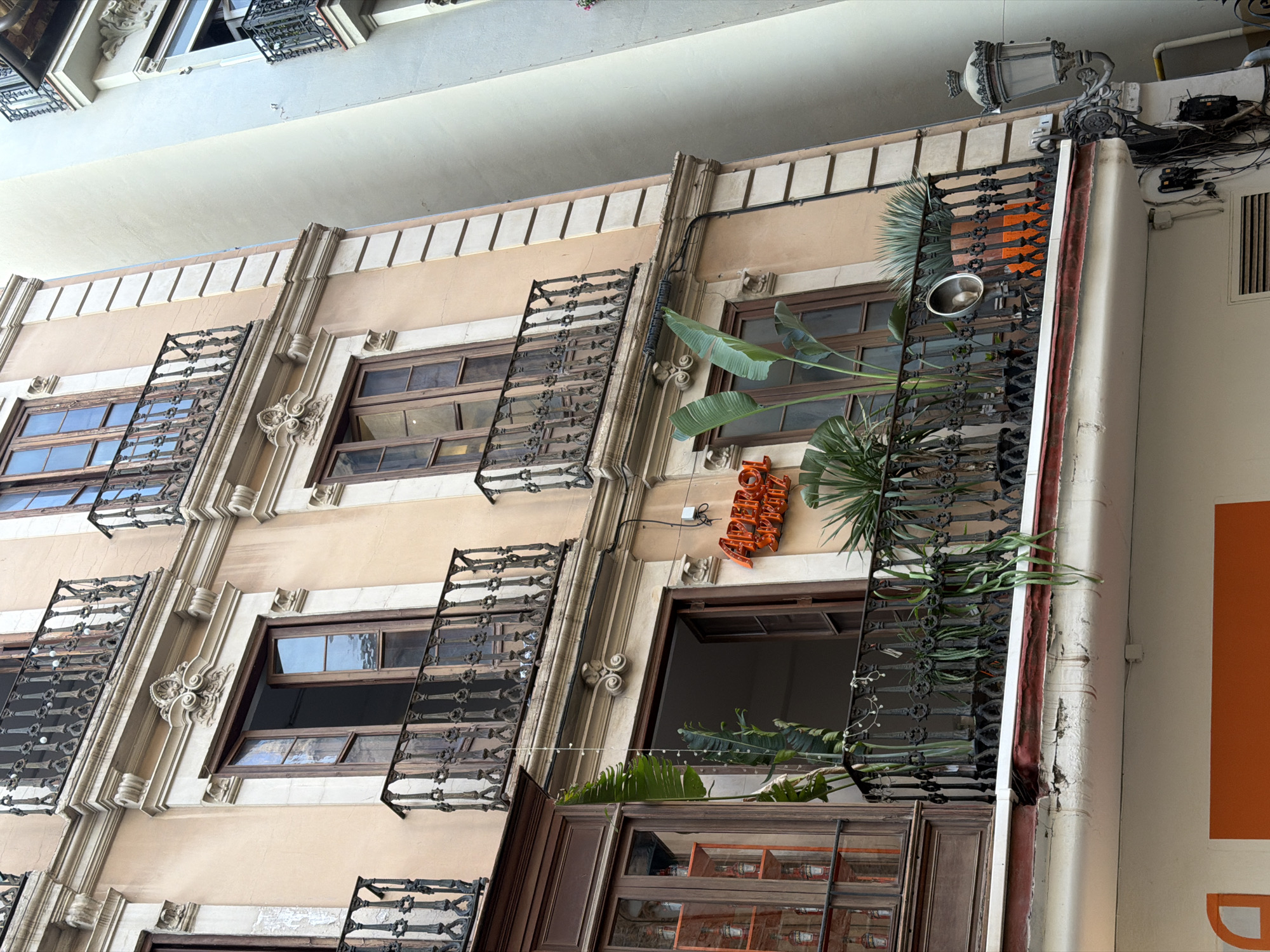 The image shows a close-up of the facade of a building in an urban environment. The building has a light pinkish-orange colored exterior with decorative architectural elements. There are multiple balconies with ornate black wrought-iron railings and wooden window frames. Some windows are open, revealing glimpses of the interior. Various plants are placed on the balconies, adding greenery to the scene. An illuminated sign with orange neon letters that reads "APEROL SPRITZ" is visible on the facade. A vintage-style street lamp is attached to the side of the building. Adjacent to the main building, another building with a pale yellow facade and a light blue facade can be seen. The image focuses on the details of the building's structure, balconies, and a prominent advertisement.
