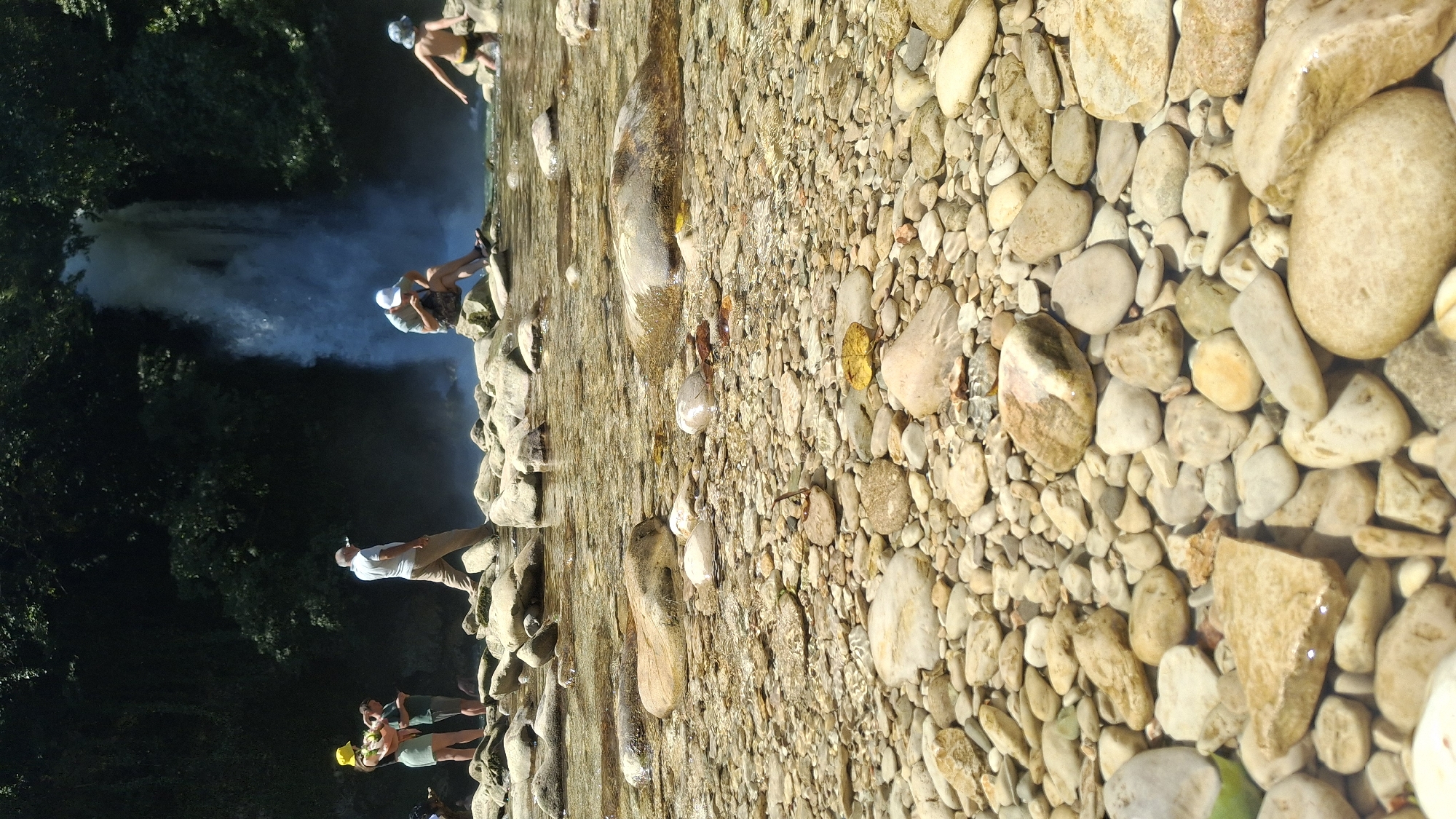 In the city of Gachedili, Georgia, a picturesque natural scene unfolds by a waterfall. In the foreground, the clear, shallow waters of a river reveal a bed of smooth, rounded pebbles and larger rocks, some submerged and others partially exposed. Sunlight filters through the water, creating shimmering reflections on the riverbed.
Further back, a majestic waterfall cascades down a rocky cliff face, partially obscured by mist. The surrounding area is densely covered with lush green foliage, typical of a forested environment.
Several individuals are present, enjoying the natural surroundings. On the left, a group of three people stand near the water's edge. One person wears a bright yellow hat and a light-colored skirt, holding a drink. Beside them, another individual in a dark shirt and shorts looks on. A third person in a green shirt and shorts stands slightly behind.
To the right of the waterfall, a man wearing a white cap sits on a rock, looking down at his phone. Nearby, a child in a blue hat and yellow swim trunks stands in the water, appearing to interact with the stream. Another person, partially visible, is further to the right, by the water. A man in a light-colored shirt and pants walks across the rocky terrain near the water's edge. The overall impression is one of a serene and refreshing natural retreat.