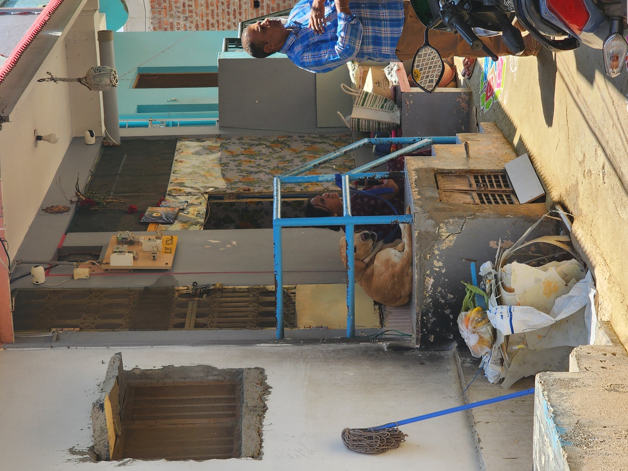 This image depicts an outdoor scene in front of a residential structure in Pedda Kammavaripalli, India.
A light-skinned, possibly concrete, building with signs of weathering and irregular plasterwork occupies the left side of the frame. An open, barred window is visible on the left wall. A blue mop leans against this wall near the bottom left corner. Adjacent to the building, a set of concrete steps leads up to an elevated entrance. The steps are worn, with chipping paint and exposed concrete.
On the elevated concrete platform, a light brown dog is curled up and resting, positioned behind a blue metal railing that acts as a banister. To the right of the dog, a woman with dark hair, wearing a patterned garment, is seated on the platform, looking towards the right side of the frame. Her right arm is visible. Behind her, the doorway is partially obscured by a patterned fabric or screen. An electrical meter box and associated wiring are mounted on the wall to the left of the doorway. Above the entrance, a light bulb hangs from the ceiling, and a decorative object resembling an ornate metal vessel is suspended by chains.
To the right of the platform, a man with short dark hair, wearing a blue and white plaid shirt and brown trousers, stands facing generally to the right, with his head turned slightly left. His right hand is raised to his chest area. He appears to be speaking or observing something outside the frame to the right. Behind him, part of a light blue building with a dark brown door is visible, along with a reddish-brown brick wall.
At the bottom of the steps, directly in front of the platform, a pile of discarded items, including plastic bags, paper, and what appears to be vegetable waste, is accumulated. On the ground level to the right, a motorcycle's front section, including handlebars, a rearview mirror with a checkered pattern, and a red taillight reflector, is visible. The ground is a mix of dirt and concrete, showing a strong shadow cast from an unseen object or building. A colorful rangoli or kolam pattern is visible on the ground beneath the motorcycle's front wheel, extending towards the building.
