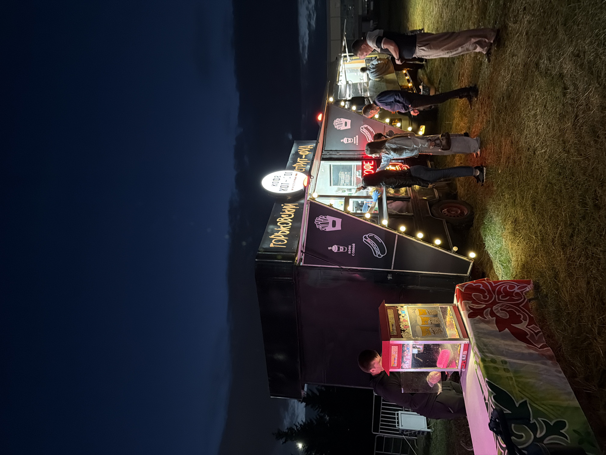 In the city of Unknown, Unknown, at dusk, a brightly lit food truck called "Gorky Street Food" is open for business. Several people are gathered around the truck, some ordering food and others waiting in line. A person in a black hoodie is attending to a popcorn machine on a table in the foreground. The food truck is decorated with lights and offers a variety of food items, including hot dogs and coffee. The background shows trees and a dark, cloudy sky.