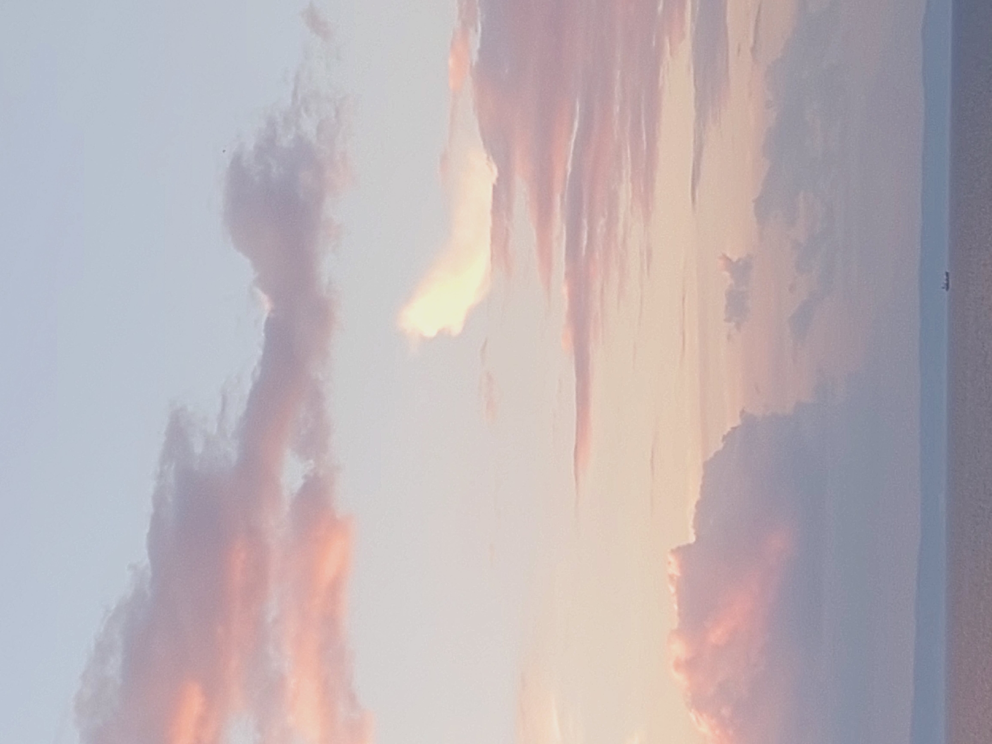 The image depicts a serene seascape at dusk or dawn, with a pastel-colored sky filled with soft clouds tinged with pink and orange hues. The sea below is calm and reflects the muted tones of the sky. In the distance, a faint, dark line suggests a landmass. A small, dark silhouette of a ship can be seen on the horizon, indicating it is sailing on the water. The overall atmosphere is tranquil and picturesque. There is no visible indication of the city of Sozopol, Bulgaria, within the frame of this image.