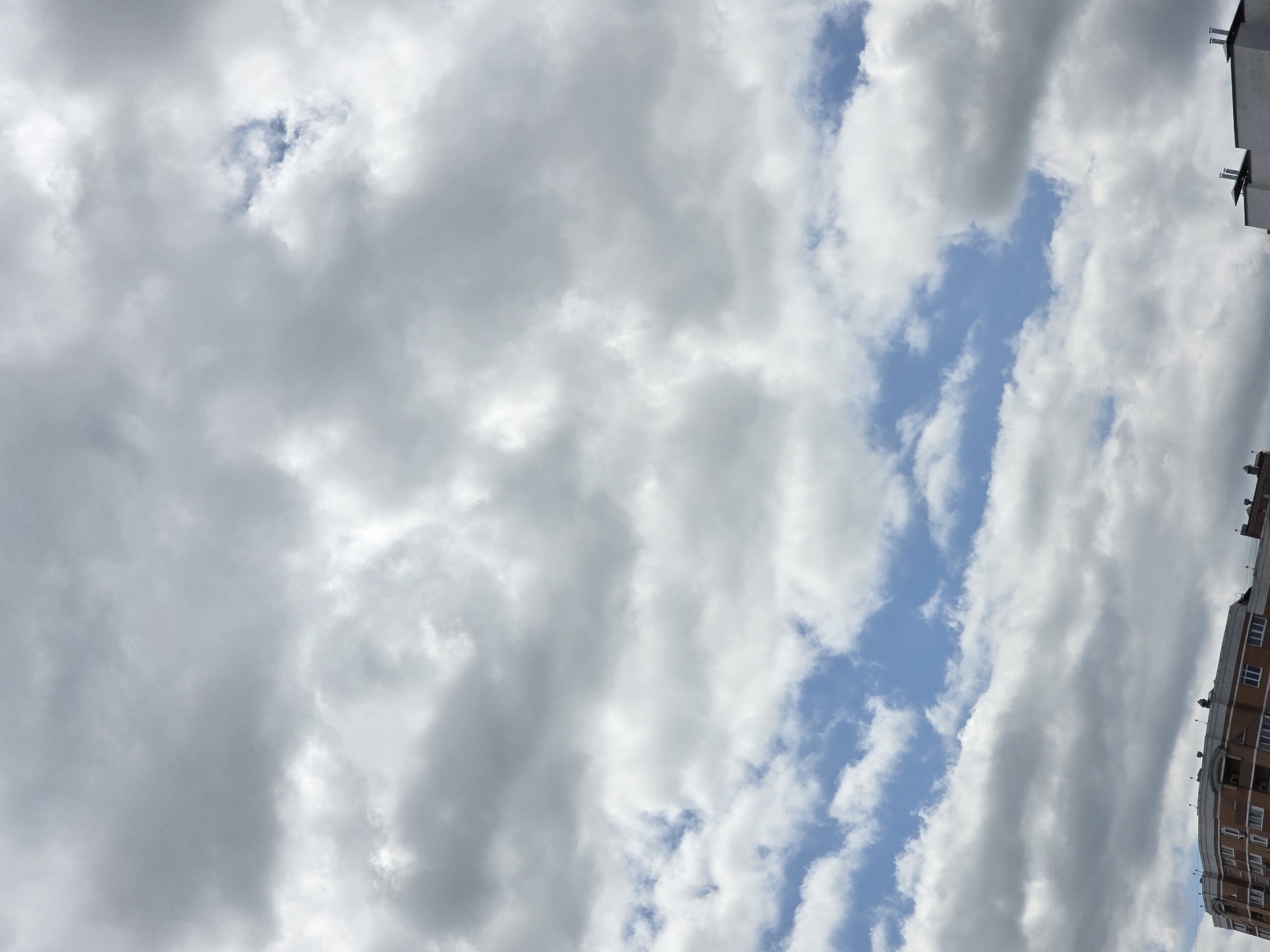 An outdoor, vertical, full shot photograph captures a sky filled with dramatic, heavy grey clouds. Interspersed within the cloud cover are patches of bright blue sky. In the bottom section of the image, the upper floors of two brick apartment buildings are visible. The building on the left appears to be older, with visible window frames and balconies. The building on the right is a lighter color, possibly white or light grey, with several protruding chimneys on its roof. The scene is devoid of any people or visible actions. The photograph was taken in Sofiivska Borshchahivka, Ukraine.