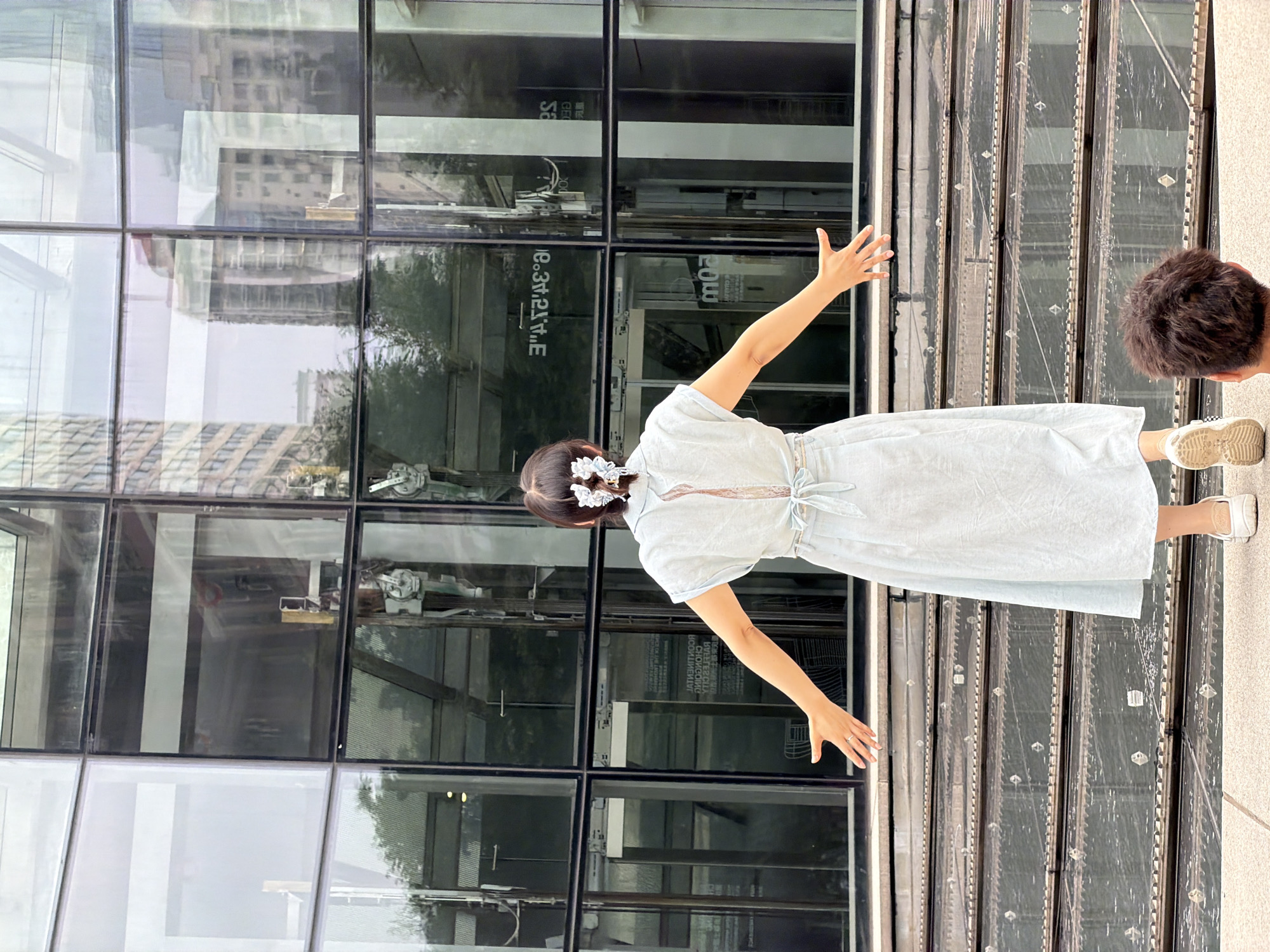 An overhead shot shows a woman in a light blue dress standing on stairs outside a modern glass building in Yuzhong District, China. She is facing away from the camera, with her arms outstretched. Her dark hair is pulled back and adorned with a white flower clip. The building's facade is made of large glass panels, reflecting the surrounding cityscape and sky. In the foreground, the back of a person's head is visible. The stairs are made of concrete with metal detailing.