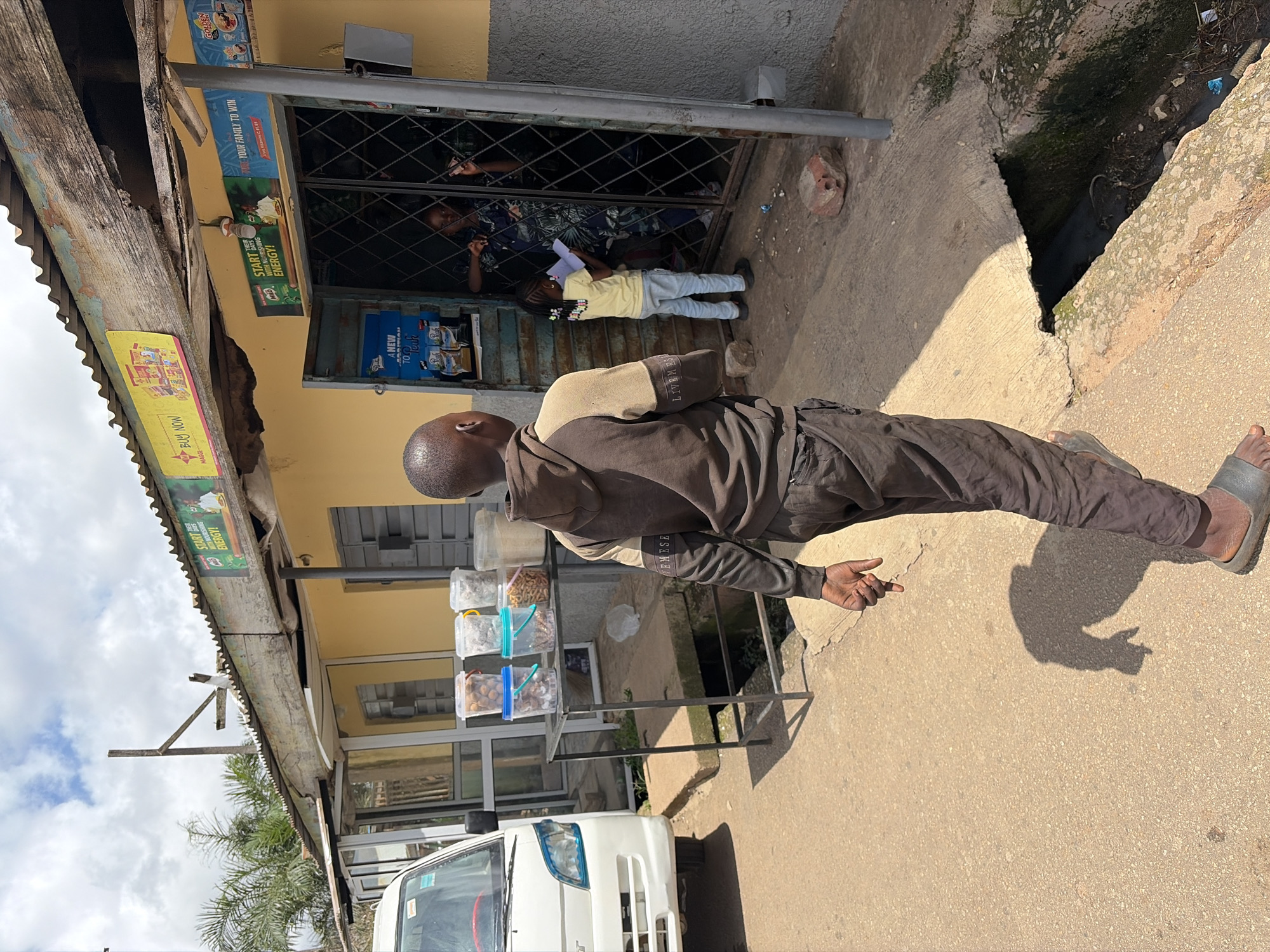 The image depicts a street scene in Jos, Nigeria. A young boy, wearing a brown and tan hooded sweatshirt and dark pants, walks away from the camera towards a small shop. The shop has a yellow facade and a corrugated metal roof with some signs advertising "MAGGI" and "MILO". In the doorway of the shop, a woman is visible, and a young girl in a yellow t-shirt and blue jeans is standing and looking at something in her hands. Outside the shop, a metal shelf holds several clear plastic containers filled with various items, possibly snacks or food items. To the left of the shop, a white van is parked. The ground is paved, and there is a concrete drainage ditch to the right of the pavement. The sky is partly cloudy.