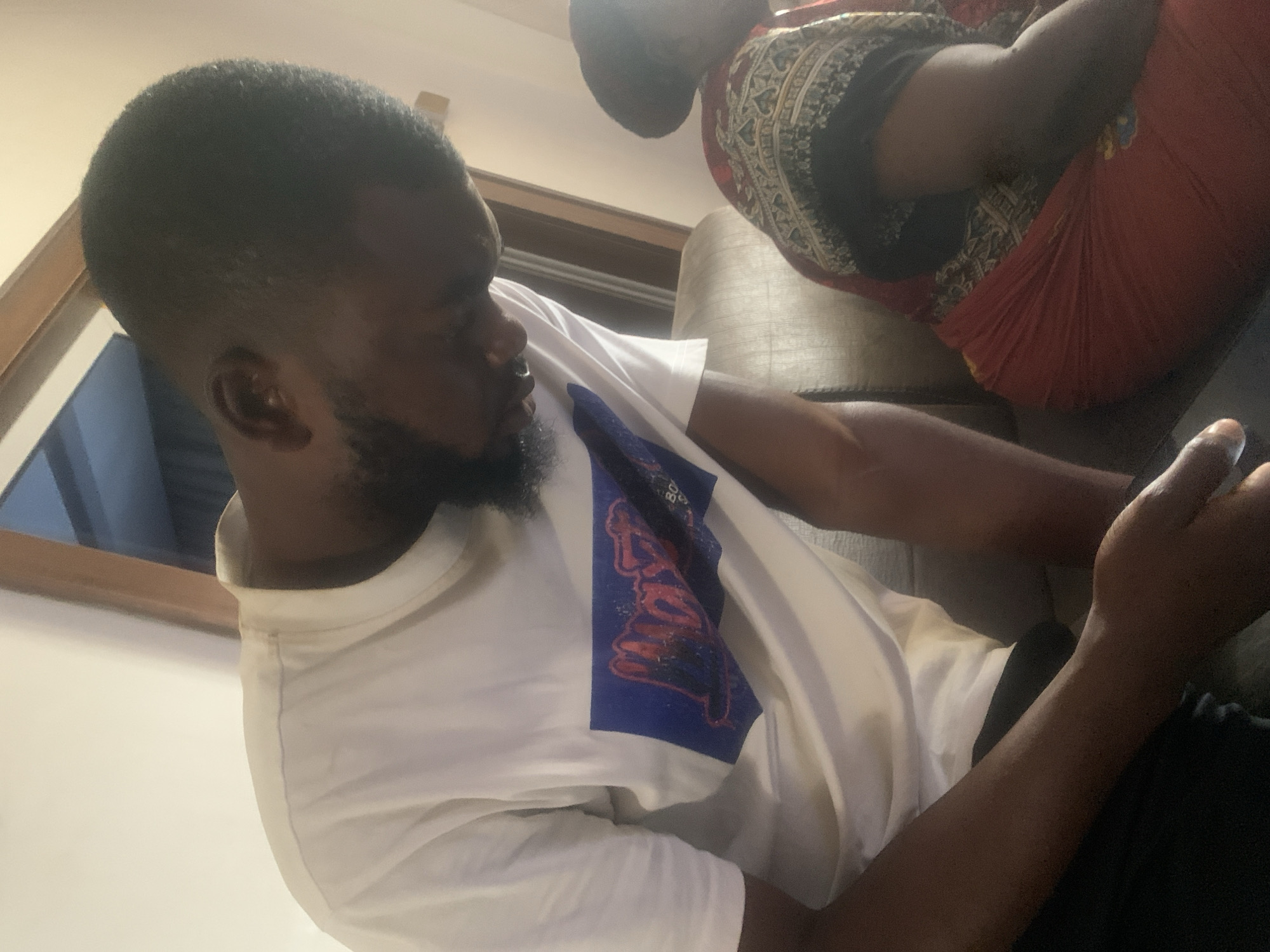 The image shows two individuals indoors, likely in a relaxed domestic setting in Jos, Nigeria. In the foreground, a man with short, dark hair and a beard is seated, turned slightly to his right. He is wearing a white t-shirt that features a prominent blue graphic design on the chest, with stylized red and white text that appears to read "BOYS." He also wears dark trousers. His left arm is bent, and his hand is raised, possibly holding an object or device that is out of frame or obscured.
In the upper right background, another person is partially visible, seated on a dark-colored couch. This individual is wearing traditional attire, characterized by a vibrant red fabric draped around them, along with an underlying garment featuring intricate black and white or cream patterns. The setting suggests an indoor space, possibly a living room, with artificial lighting casting a warm glow. A wooden structure, perhaps part of a furniture piece or interior decor, is visible behind the man. The scene conveys a casual and quiet moment.