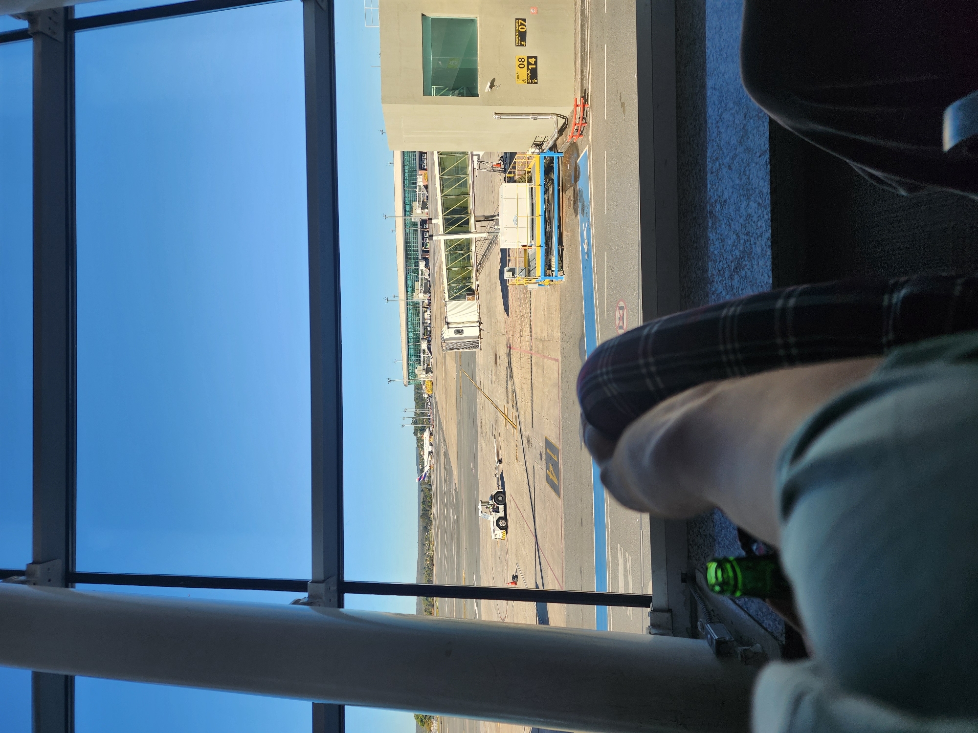 This media file shows an airport scene viewed from inside through a large window. The sky is clear and blue, suggesting a sunny day. The time of day appears to be daytime, likely late morning or early afternoon due to the strong sunlight casting shadows.
The scene depicts an airport tarmac with several elements visible. A jet bridge connects to an airport terminal building, which has several windows and signage. Various vehicles, including baggage carts and a pushback tractor, are present on the tarmac. In the distance, multiple aircraft can be seen parked at gates or on runways. The overall setting is an active airport.
In the foreground, parts of a person are visible. This includes their legs, one wearing plaid pants, and an arm holding a green glass bottle. The person appears to be relaxed, possibly waiting for a flight. The angle of the shot and the presence of the person's belongings suggest they are seated near the window.
The location is identified as Lago Sul, Brazil. There are some visible text elements, including numbers on the tarmac that appear to be gate or parking bay indicators, such as "14". There is also some text on the terminal building that is partially obscured and difficult to read clearly. The overall mood conveyed is one of waiting and observation within an airport environment.