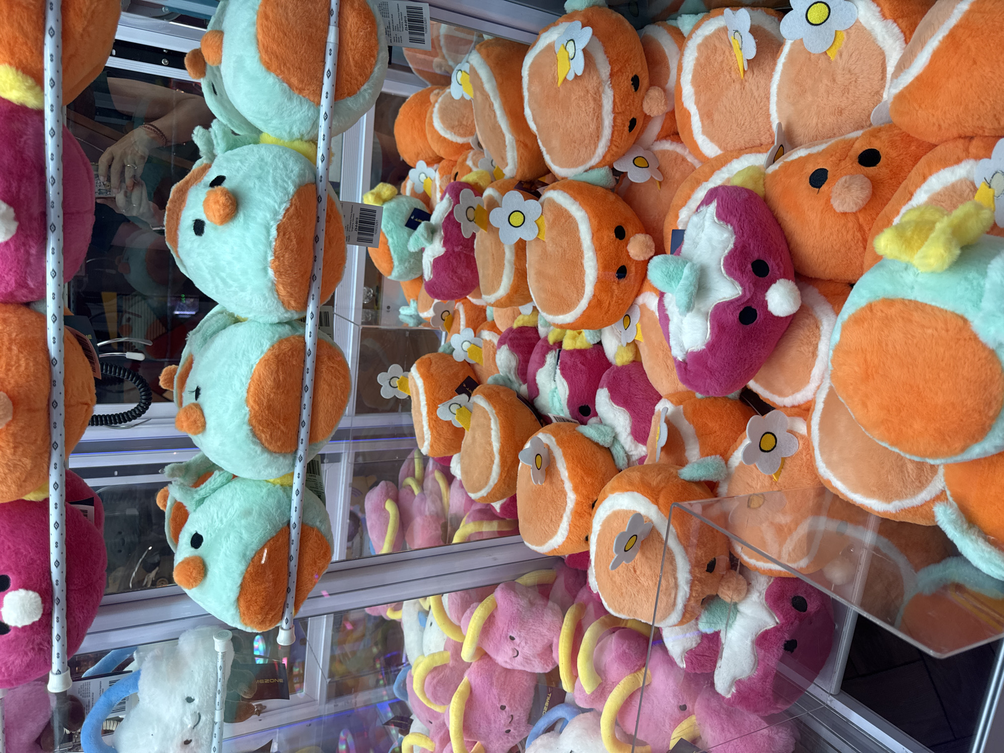 The image shows a claw machine filled with plush toys. The toys are arranged in multiple rows. The primary type of plush toy is circular, with a white outer ring and a flower detail. These toys are mostly orange, with some being teal and pink. Below these are pink plush toys with yellow appendages. The location is not specified, and cannot be inferred from this image.