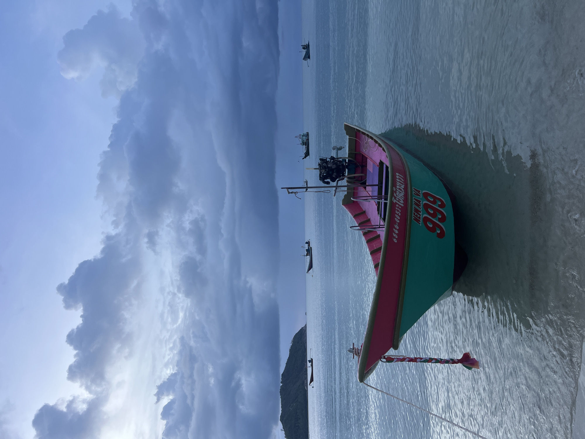 A fishing boat with a turquoise hull and a pink interior is anchored in shallow, clear water near a sandy beach. The boat has a small outboard motor mounted at the stern and is tethered by a rope to the shore. The number "999" is painted in red on the boat's hull. In the background, several other fishing boats are visible on the ocean, and a lush green island with a hilly terrain is situated on the horizon. The sky is overcast with dramatic, dark clouds, suggesting either dawn or dusk. The scene is set in the Ko Pha-ngan District of Thailand.