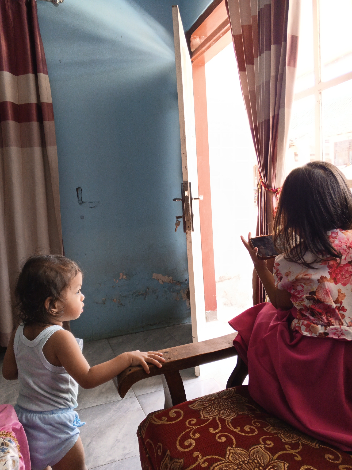 In Singolatren, Indonesia, two young girls are present in a room with blue walls and a light-colored tiled floor. A doorway with a door ajar reveals a brightly lit outdoor area. The doorway is equipped with a handle and a hanging key. To the left, a curtain with maroon and beige stripes hangs. A second curtain, also with maroon and beige stripes, is partially visible near the doorway.
The younger girl, facing left, wears a white tank top and blue shorts. She stands next to a wooden chair and touches its side. The older girl, seen from the back, is seated on a chair covered with a red and gold patterned fabric. She wears a floral-print top and a magenta skirt. She is holding a smartphone horizontally, seemingly engrossed in its screen.