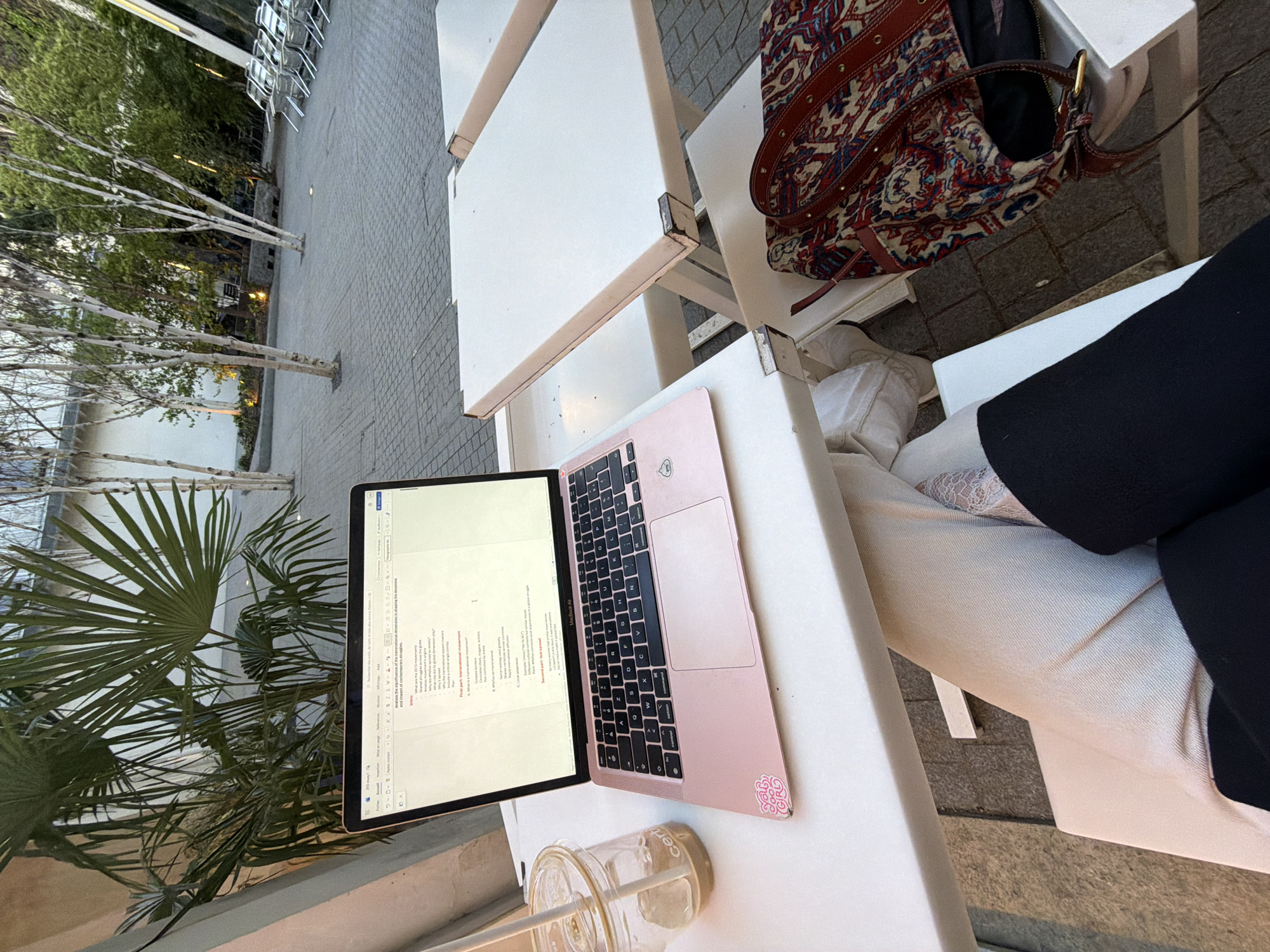 The image depicts a first-person perspective of a person seated outdoors at a white table in Paris, France, engaged in work on a laptop.
In the immediate foreground, the individual's lower legs are visible, dressed in light-colored trousers or a skirt, with a dark garment draped over the lap. Their right foot shows a light-colored, possibly lace, sock or shoe.
On the white table directly in front, a pink MacBook Air laptop is open, displaying a document on its screen. The document, shown in a word processor interface with French menu options ("Fichier," "Accueil"), contains English text related to "transnational movement," "2015 movements," and "shared struggles." A pink sticker, likely "GRL PWR," is affixed to the laptop's left palm rest. To the left of the laptop, a clear cup with a straw holds an iced beverage, with the partial word "CERC" or "CERT" visible on its side.
To the right of the laptop, another section of the white table is visible, showing signs of wear on its edges. An adjacent white chair supports a red and multi-colored patterned shoulder bag, adorned with leather straps and metal studs.
The background reveals a spacious, paved outdoor area composed of gray square pavers. Several slender, white-barked trees (birch trees) are scattered across this paved surface. Behind these trees, dense green foliage suggests a garden or park-like setting, with glimpses of distant outdoor chairs and subtle artificial lighting elements near the ground. A large, fan-leaved palm-like plant is situated immediately behind the laptop, contributing to the outdoor ambiance. The overall environment suggests a modern, well-maintained outdoor public space, such as a café terrace, a university campus courtyard, or a cultural institution's plaza in Paris.