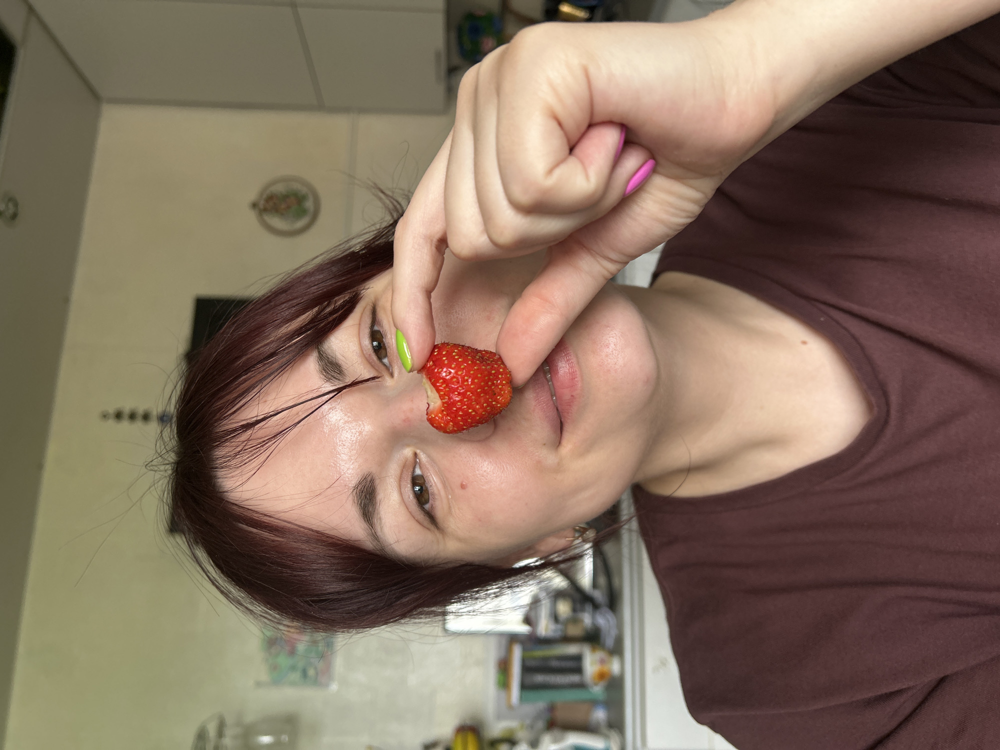 The media file shows a close-up, rotated portrait of a person holding a strawberry near their nose. The person has dark red hair and brown eyes, with a light complexion. Their fingernails are painted in bright colors: pink on the right hand and lime green on the left. The strawberry is red and appears to be fresh. The person is wearing a dark maroon t-shirt.
The background suggests an indoor setting, possibly a kitchen, with cabinets visible in the upper left corner. There are some out-of-focus objects in the background, including what might be shelves with various items. The lighting appears to be artificial, typical of an indoor environment.
The activity is simple: the person is posing with a strawberry, perhaps for a playful selfie or as part of a creative shot. There are no strong emotional cues, but the pose might suggest a lighthearted or whimsical mood. No visible text is present in the image. The location context provided is "Фоки, Russia," but there are no specific visual cues within the image that directly confirm this location.