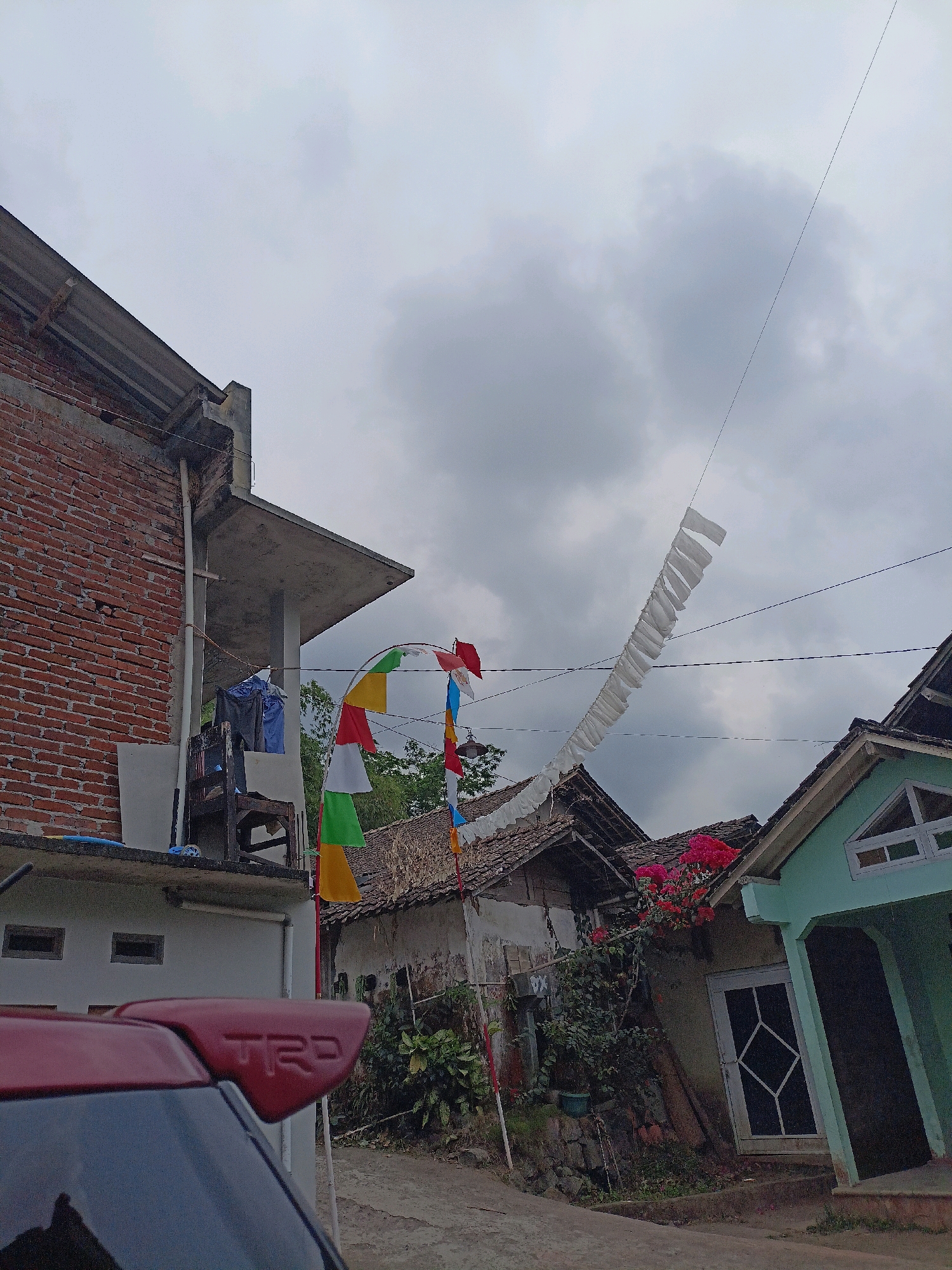The image shows a street scene in Ngepanrejo, Indonesia. The sky is overcast with dark clouds. On the left, a brick building with a concrete overhang extends upward. A white pipe runs vertically down the side of the building. Below the overhang, a wooden chair with laundry hanging on it is visible. Several colorful triangular flags are strung across the scene, along with a long string of white rectangular flags.
In the mid-ground, there are several traditional Indonesian houses with tiled roofs. One house on the right is painted light green and has a white-framed door and window. Red bougainvillea flowers are visible on the side of this house. A red car with a TRD spoiler is partially visible in the foreground on the left. The street is unpaved.