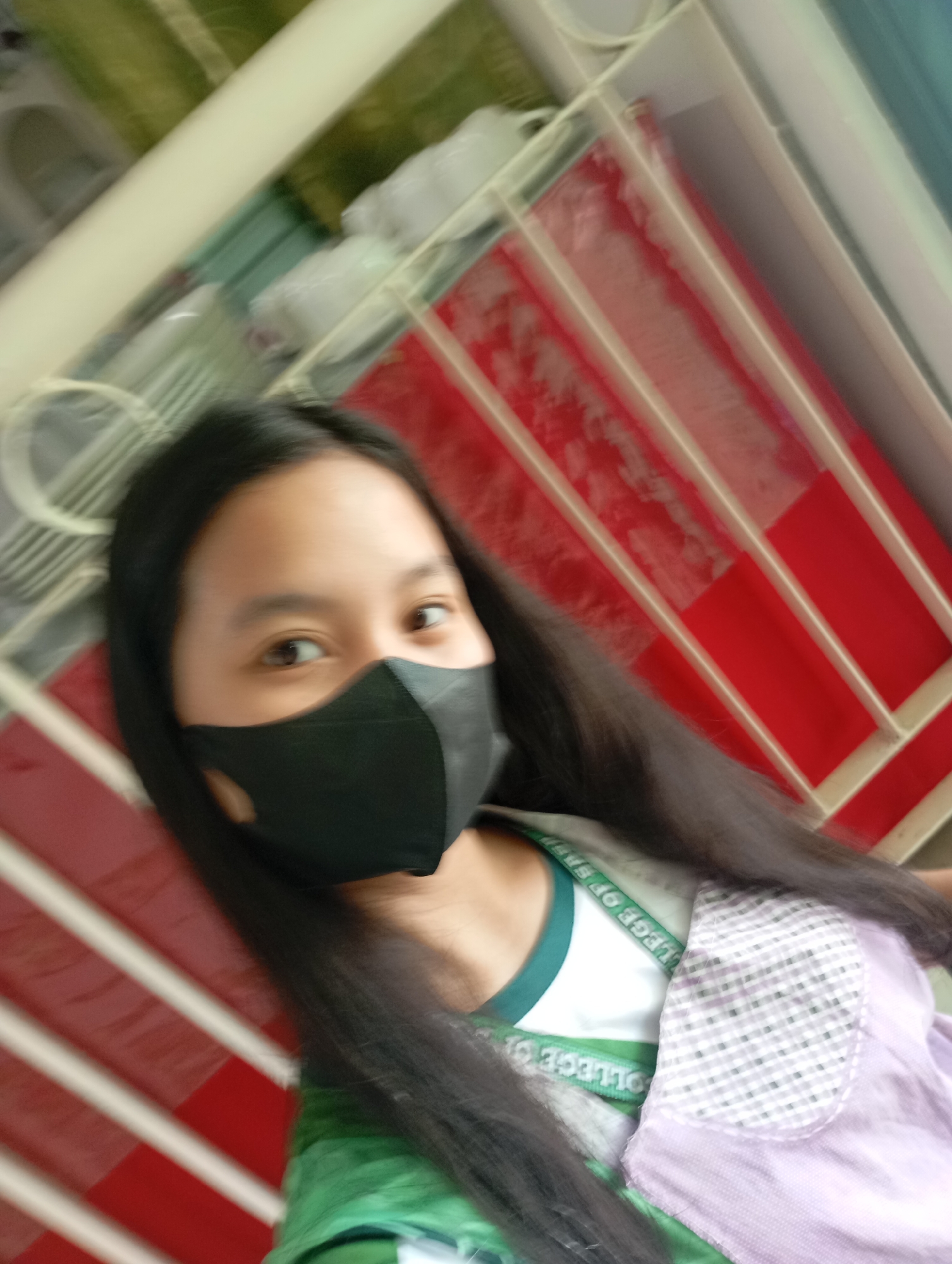 A slightly blurry, diagonally oriented image captures a close-up portrait of a young person, likely female, wearing a black face mask. Her long, dark hair frames her face, with one eye visible and looking towards the viewer. She is wearing a green and white top, possibly a uniform, with a visible strap or collar that features partially legible text: "COLLEGE OF SP..." is discernible, repeated and reversed on the strap. Over this, a light purple or lavender garment with a small, checkered-patterned flap or pocket is worn. The background is heavily blurred but appears to be an indoor or semi-enclosed setting, featuring white diagonal bars against patches of red and green, possibly a shelving unit, railing, or architectural detail. The lighting suggests daytime. The overall impression is a casual selfie, potentially taken in Davao City, Philippines, with motion blur or shallow depth of field contributing to the aesthetic.