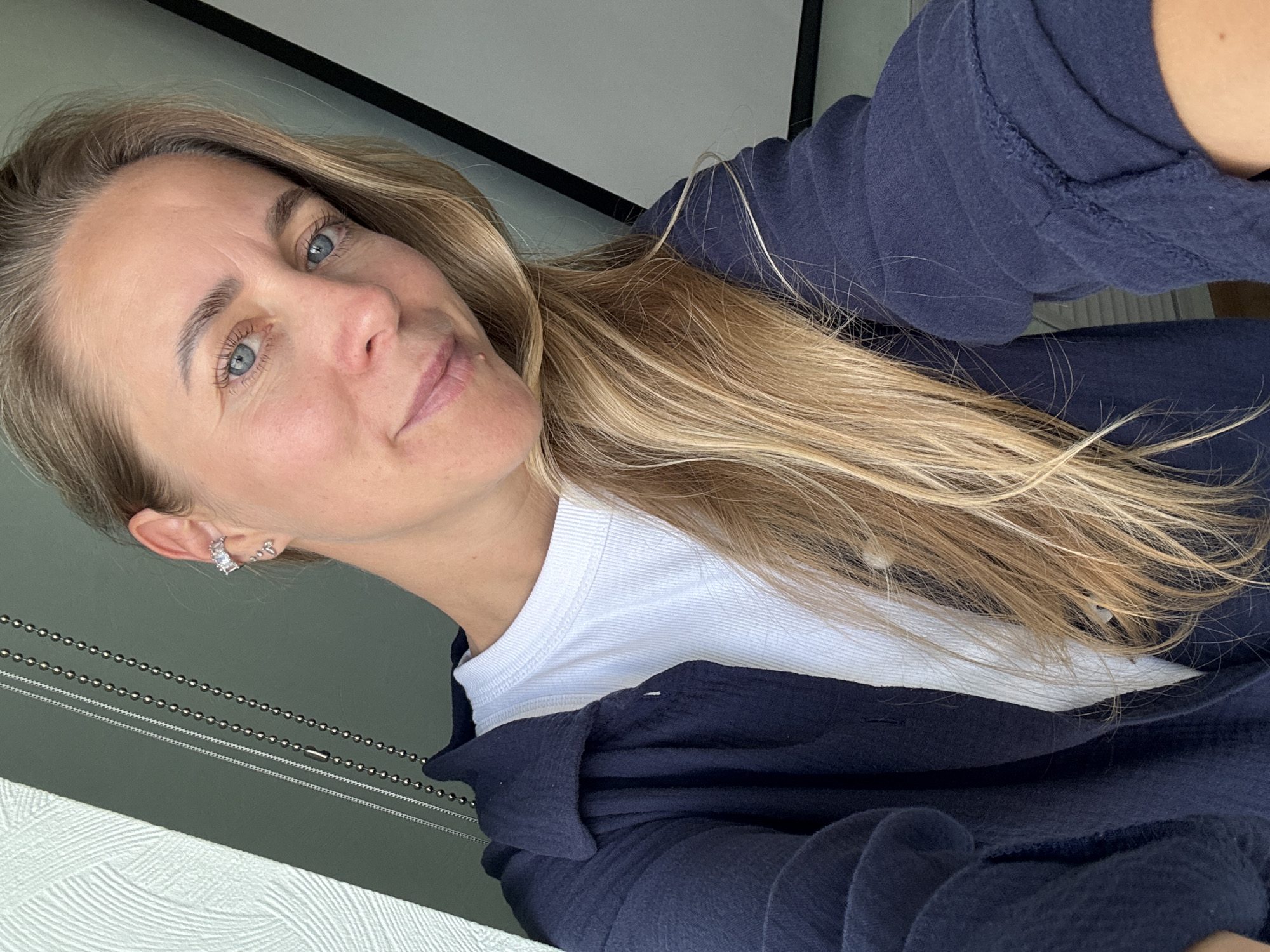 A woman with blonde hair and blue eyes takes a selfie. She is wearing a white t-shirt and a dark blue shirt. She has a silver ear cuff with three stones on her left ear. Her hair is long and blonde, falling over her right shoulder. The background is blurred and appears to be an indoor setting with a window on the left and a white projection screen on the right. The lighting suggests it is daytime.