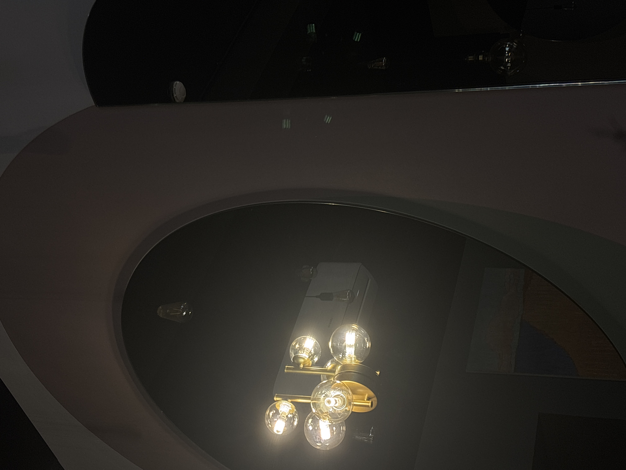 The image captures a contemporary interior scene, likely at night, within the context of Perm, Russia, characterized by artificial lighting and reflective surfaces. The main subject is a modern chandelier, composed of a golden or brass-toned fixture holding several clear, spherical light bulbs, which are illuminated and emit a warm glow.
This chandelier is prominently reflected in a large, dark, oval-shaped surface, which appears to be a polished ceiling panel or a decorative mirror. Above this oval, another dark, curved surface reflects faint, striped patterns, suggesting another light source or textured object in the environment. Further reflections show single, exposed light bulbs. Within the large oval reflection, to the right of the main chandelier, there is a subtle reflection of abstract shapes and colors, hinting at a nearby artwork or a patterned wall section with blue, orange, and gray tones. The overall architecture features dark gray and black curved and angular elements, contributing to a sophisticated and dimly lit ambiance. No people or text are visible, and no specific activity is taking place.
