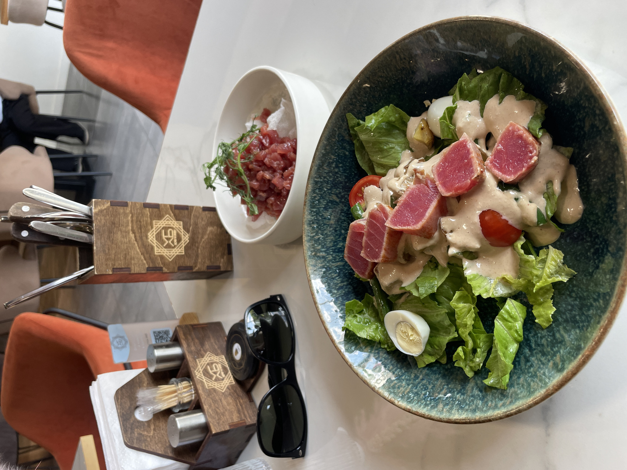This image presents a top-down view of a dining table in what appears to be a modern restaurant or cafe in Kazan, Russia. The scene primarily showcases two dishes and various table accessories, suggesting a meal is either being served or has just begun.
The main subject is a large, round, dark teal-blue ceramic bowl containing a fresh salad. The salad features crisp green lettuce leaves, vibrant red cherry tomatoes, and several slices of perfectly seared tuna, which are pink and rare in the center with a lightly cooked exterior. A creamy, light-brown dressing is generously drizzled over the ingredients, alongside a halved quail egg and other light-colored components.
Next to the main salad, a smaller white bowl holds a portion of tuna tartare, consisting of finely diced red fish topped with green microgreens and resting on a base of white, puffy crisps.
On the table, there are two distinctive wooden boxes, each adorned with a geometric logo that appears to be a stylized '9' and 'X' or similar design within a square. The larger box serves as a cutlery holder, filled with various forks and knives featuring dark handles. The smaller box is a condiment holder, containing two cylindrical silver shakers (likely for salt and pepper) and a section for toothpicks, with white napkins neatly stacked beside it. A pair of black Wayfarer-style sunglasses is also resting on the table.
In the background, glimpses of upholstered chairs—one in a warm orange color and another in a muted beige—are visible, contributing to the indoor restaurant setting. A blurry QR code can also be partially seen on a small card near the condiment holder. The table surface is light-colored, possibly marble or quartz. The overall impression is one of a contemporary dining experience, likely taking place during the daytime given the natural, ambient lighting.