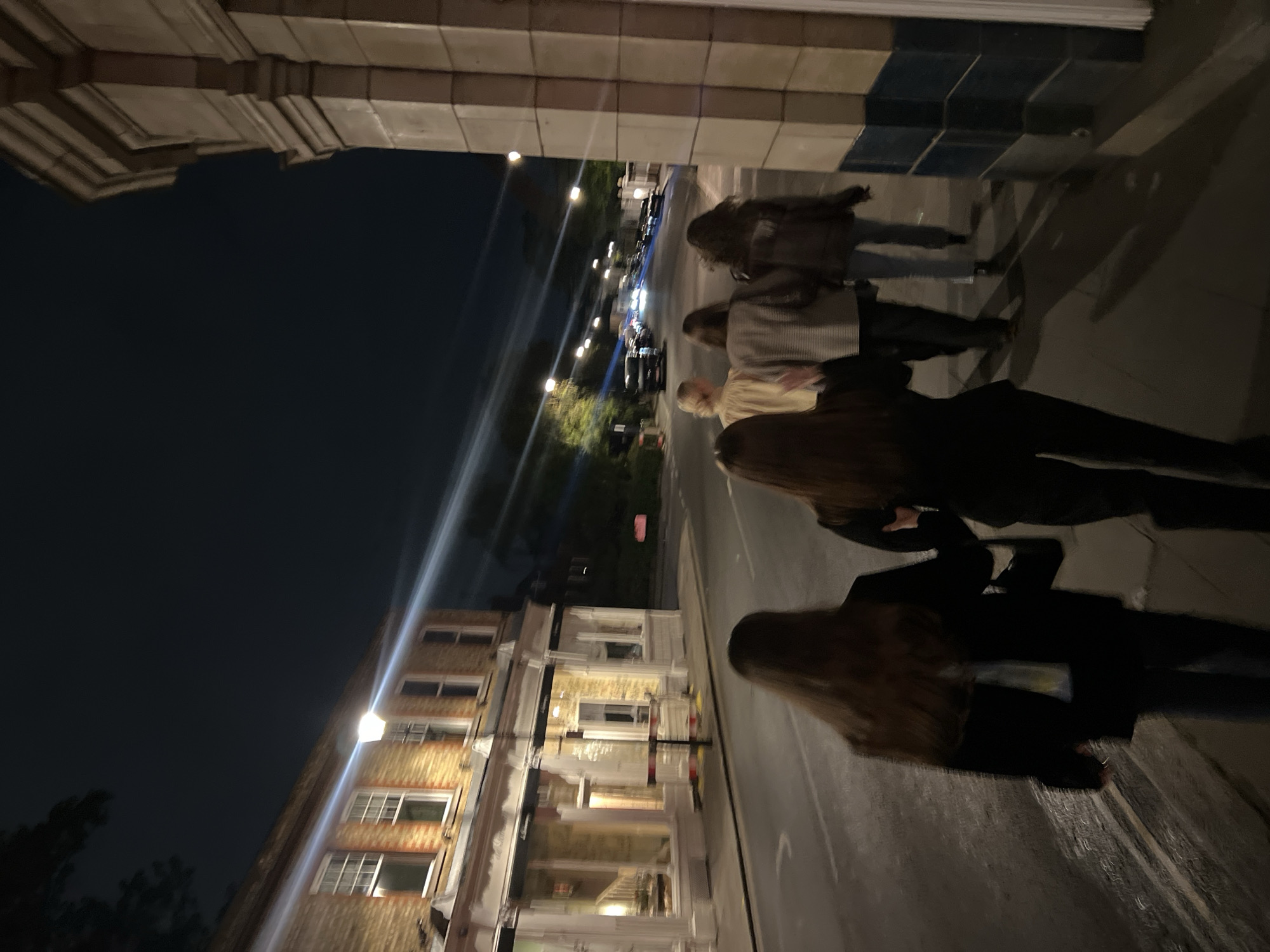 The image depicts a nighttime urban street scene, likely in London.
**Subjects:** The primary subjects are a group of several individuals, mostly seen from behind or the side, walking along what appears to be a sidewalk or pedestrian area. Their faces are obscured by low light and motion blur. They are dressed in casual outerwear, such as coats and jackets. In the background, multi-story brick buildings with numerous windows line the street. Several streetlights are visible, emitting bright light. Cars are present on the street, identifiable by their headlights and taillights, which appear as streaks due to motion blur. An architectural element, possibly an archway or ornate cornice of a building, frames the upper left portion of the image.
**Activity or event taking place:** The scene captures people walking along a city street at night, suggesting general pedestrian movement. The presence of vehicles indicates ongoing road traffic. It portrays an everyday evening atmosphere in an urban setting.
**Notable details:** The time of day is clearly night, with a dark sky and the urban environment illuminated solely by artificial light sources. The low light conditions contribute to significant motion blur, particularly evident in the moving figures and vehicle lights, creating light streaks. Bright lens flares are also visible from the streetlights. The architecture, characterized by brick buildings and window styles, is consistent with the urban landscape of London. No specific weather conditions are indicated beyond the darkness.
**Visible text:** No legible text or signage is visible in the image.