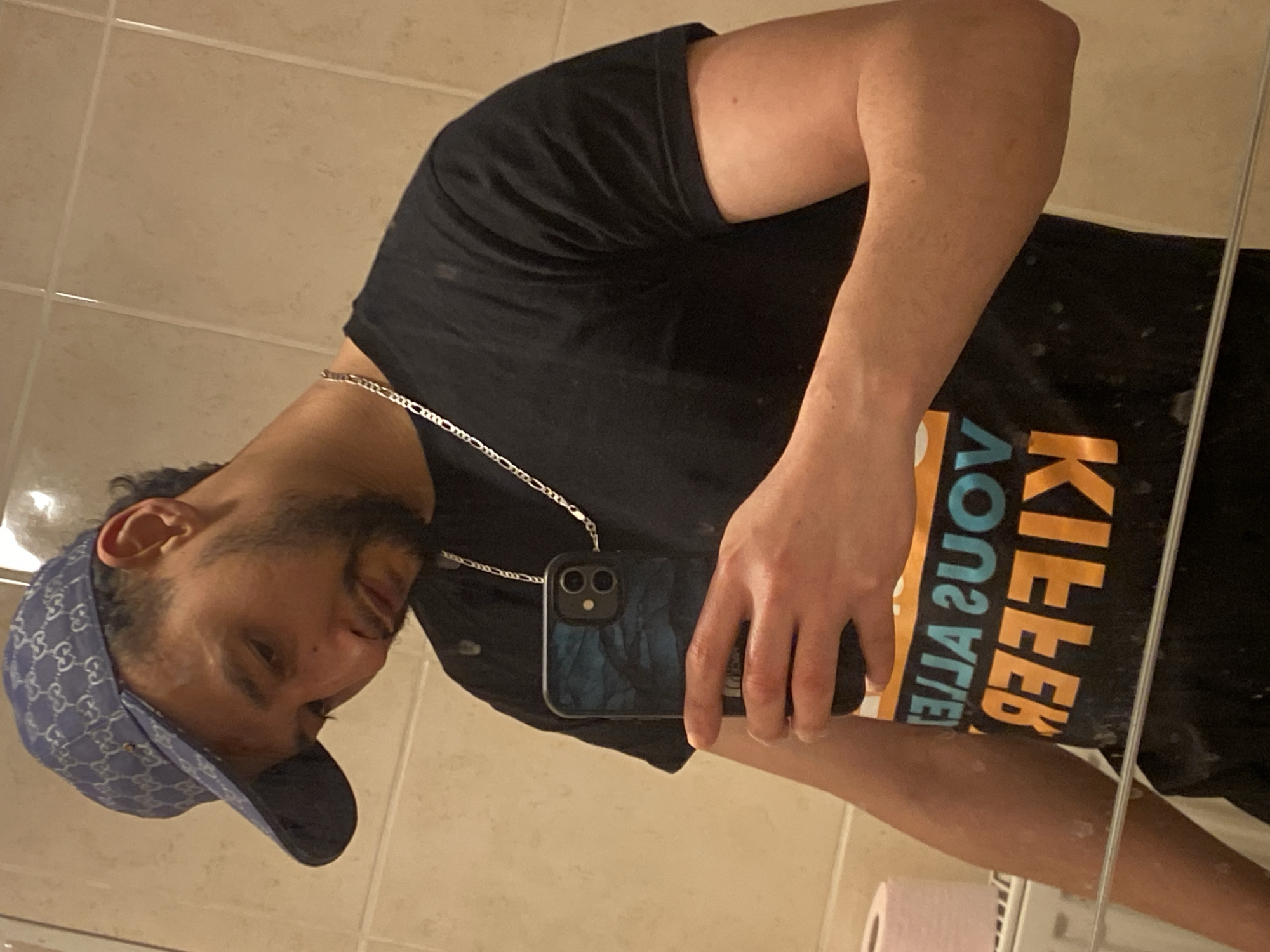 A man is taking a selfie in a bathroom. He is wearing a black t-shirt with the words "KILLER YOU ARE" printed on it, a blue Gucci baseball cap, and a silver chain necklace. He is holding a blue iPhone in his left hand. The bathroom has white tiles, a toilet, and a roll of toilet paper. There is a reflection of the man in the mirror. The location is not specified.