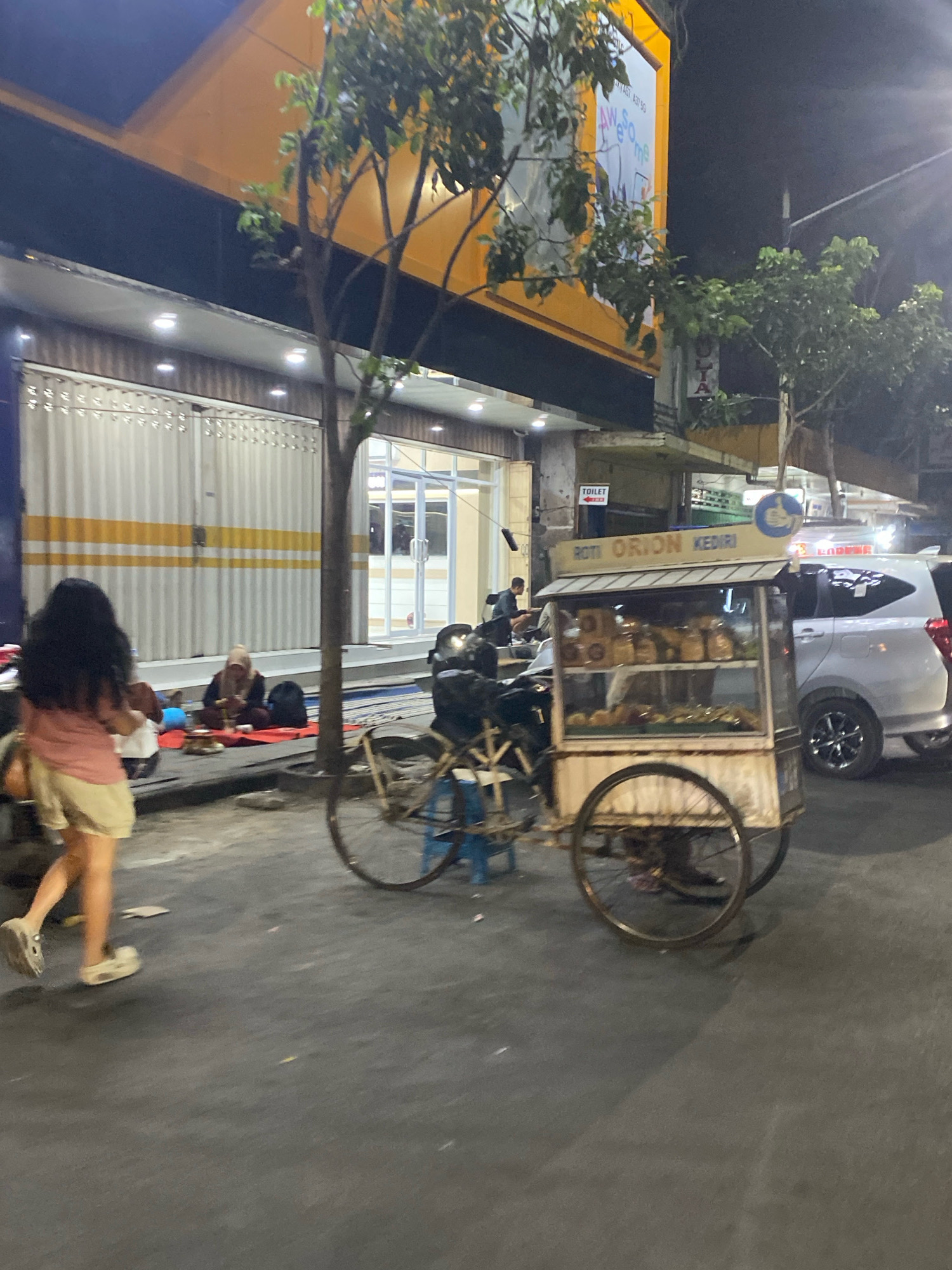 The image depicts a street scene in Kediri City, Indonesia, at night. In the foreground, a woman with long dark hair is walking away from the camera, wearing a pink shirt and beige shorts. To her left, two people are sitting on the ground, engaged in conversation or eating. A small cart selling bread, labeled "ROTI ORION KEDIRI," is visible in the middle ground. The cart has a glass display case filled with bread. Behind the cart, a silver car is parked. The background shows a building with a yellow awning and a dark facade, illuminated by lights. Trees are scattered throughout the scene. The overall environment suggests a bustling urban setting with street vendors and local residents.