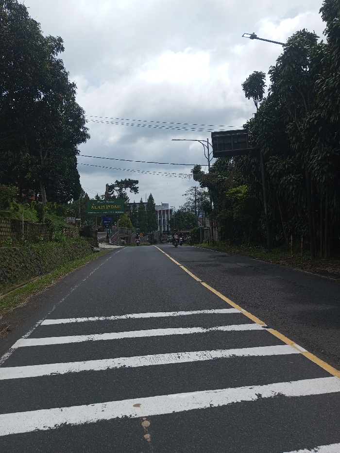 The image displays a two-lane asphalt road, viewed from a low angle, extending uphill under an overcast sky. In the immediate foreground, white pedestrian crossing stripes are painted across the road, with a solid yellow line marking the right shoulder. To the left, a sloped embankment covered in green foliage and stone retaining walls borders the road. To the right, dense green trees and bushes line the road. Further up the road, a large green overhead sign is visible, bearing the text "ALAM INDAH". This text, translated as "Beautiful Nature" or "Beautiful Realm," indicates the location is likely in an Indonesian or Malaysian speaking region, contradicting the provided location of Gubbisamamidi, India. Two motorcycles are present on the road ahead, moving away from the viewer. In the distance, beyond the sign and motorcycles, a multi-story building with light-colored facades is visible. Utility wires crisscross the upper portion of the scene, and a street light fixture extends from the right side. The overall lighting is soft and diffused due to the heavy cloud cover.