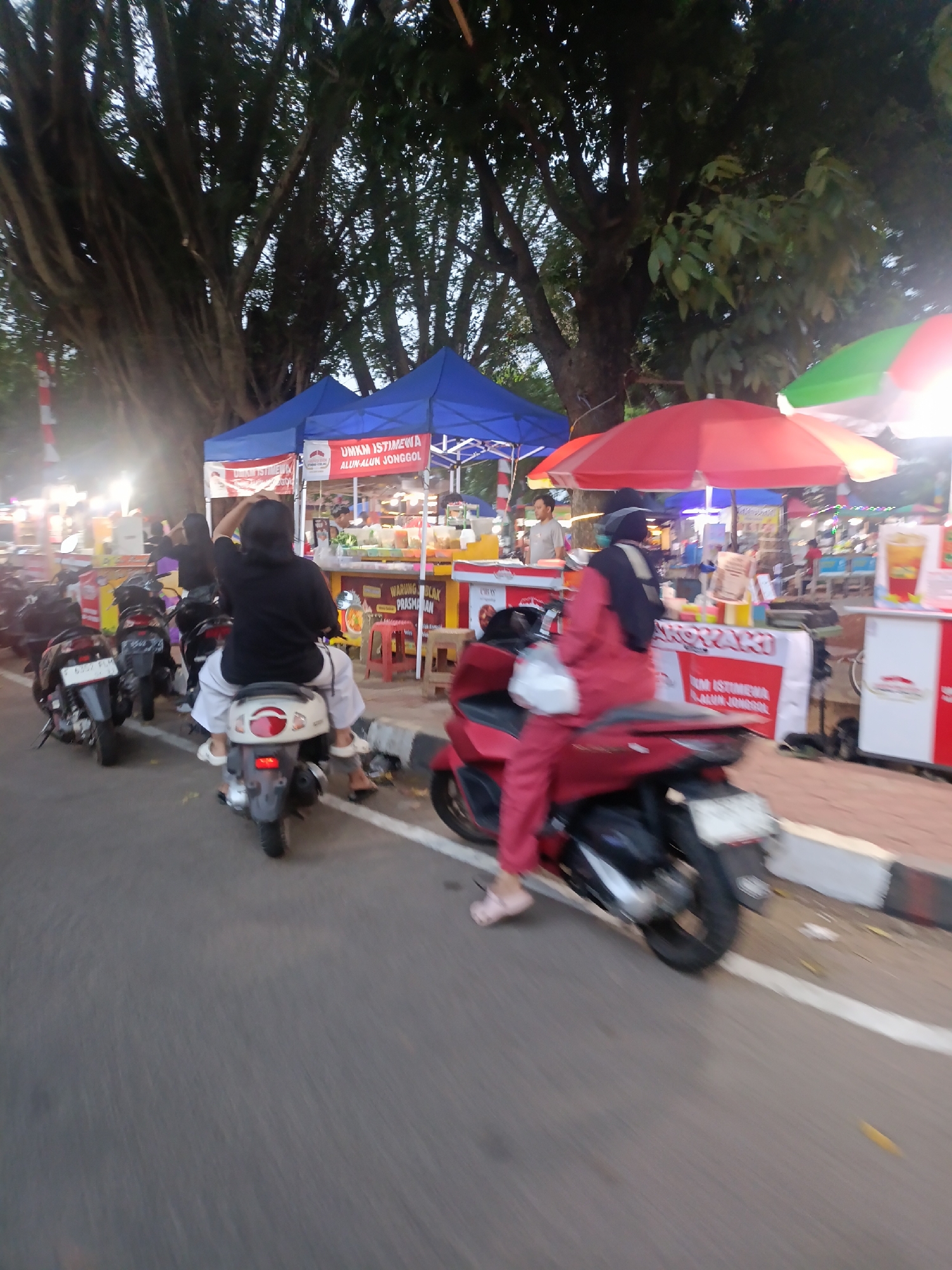 The image depicts a street scene in Jonggol, Indonesia, likely a market or event area. In the foreground, two individuals are riding motorbikes. The person on the left, wearing a black shirt and white pants, is on a light-colored scooter. The person on the right is on a red scooter, wearing a red outfit and a dark headscarf, and is looking to their left. Several other motorbikes are parked or in motion in the background.
Behind the motorbikes are several stalls or vendor booths, some with blue and red awnings. One stall has a sign that reads "UMKM ISTIMEWA ALUN-ALUN JONGGOL," indicating it's a special UMKM (Micro, Small, and Medium Enterprises) stall at the Alun-Alun (town square) of Jonggol. The stalls display various goods, and some appear to be selling food or drinks, with visible signage and products. A large tree with dense foliage dominates the upper portion of the image, providing shade. The overall lighting suggests it is either early morning or late afternoon. The ground is paved, with a curb and a white line marking the edge of the road.